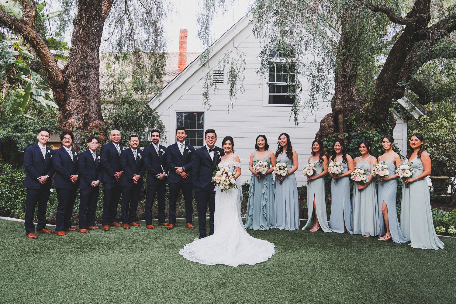 Wedding party in navy suits and light blue dresses poses together on manicured lawn in front of white building.