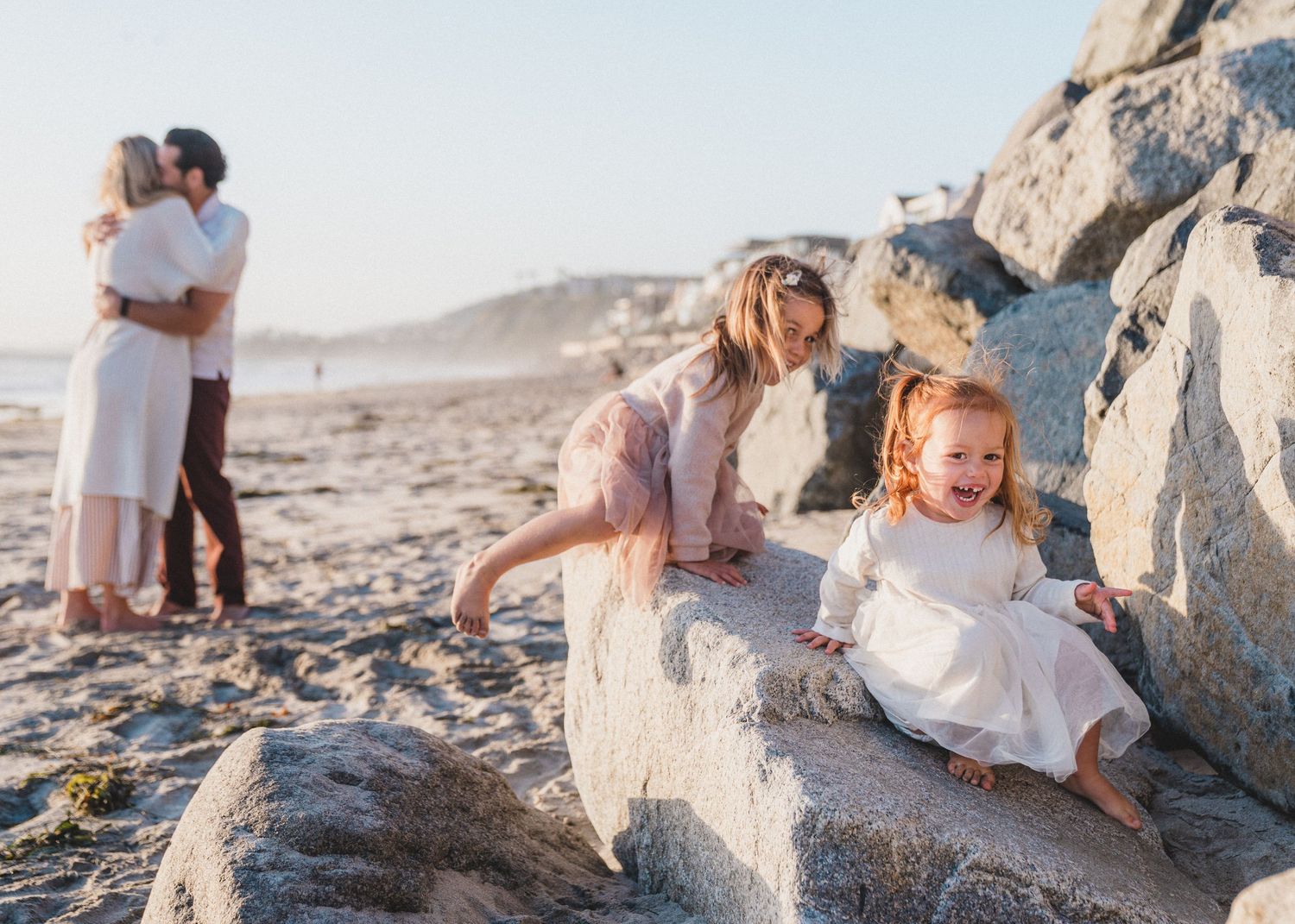 A couple in white outfits embraces on a rocky beach during sunset while having their photos taken.