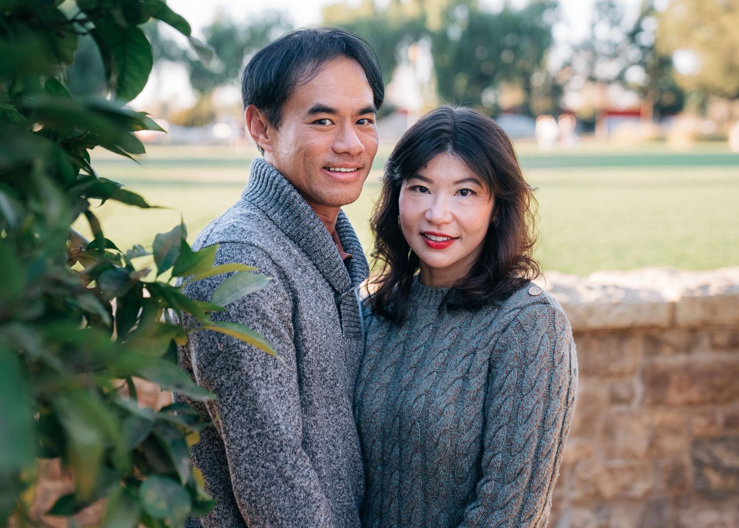 A couple in warm sweaters poses together against a stone wall with greenery in the background.