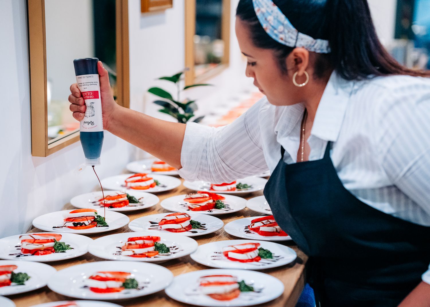 Chef in black apron garnishes multiple plates of caprese salad with balsamic glaze in restaurant kitchen.