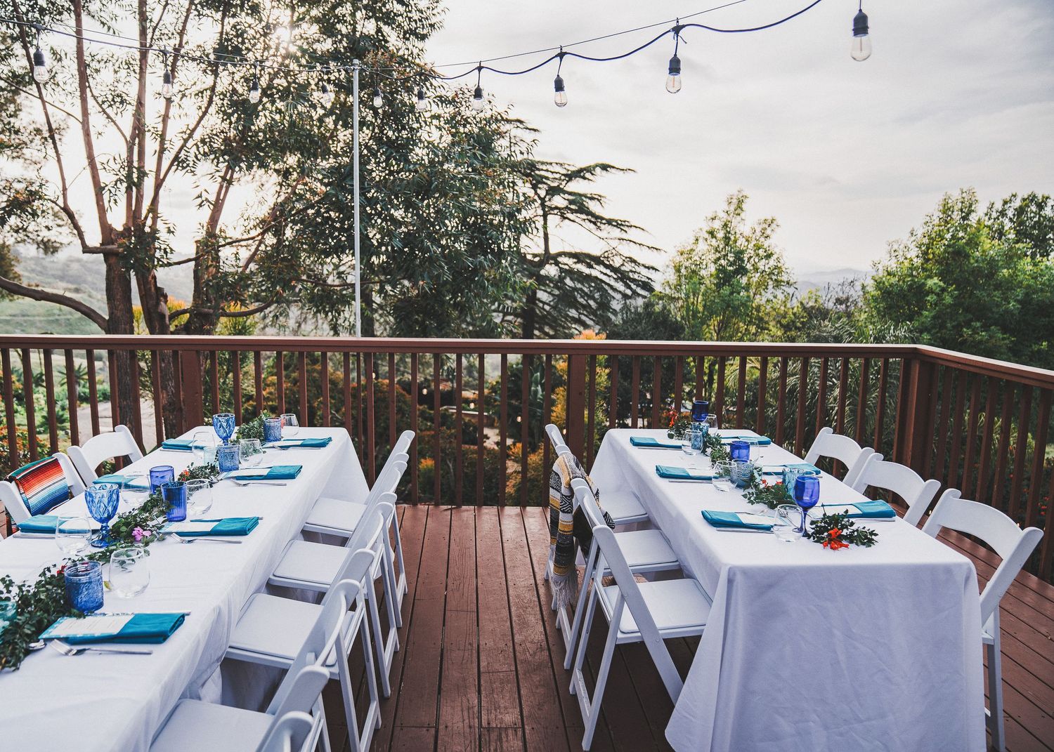 Outdoor deck reception setup with white tables and blue napkins under string lights and tall trees.