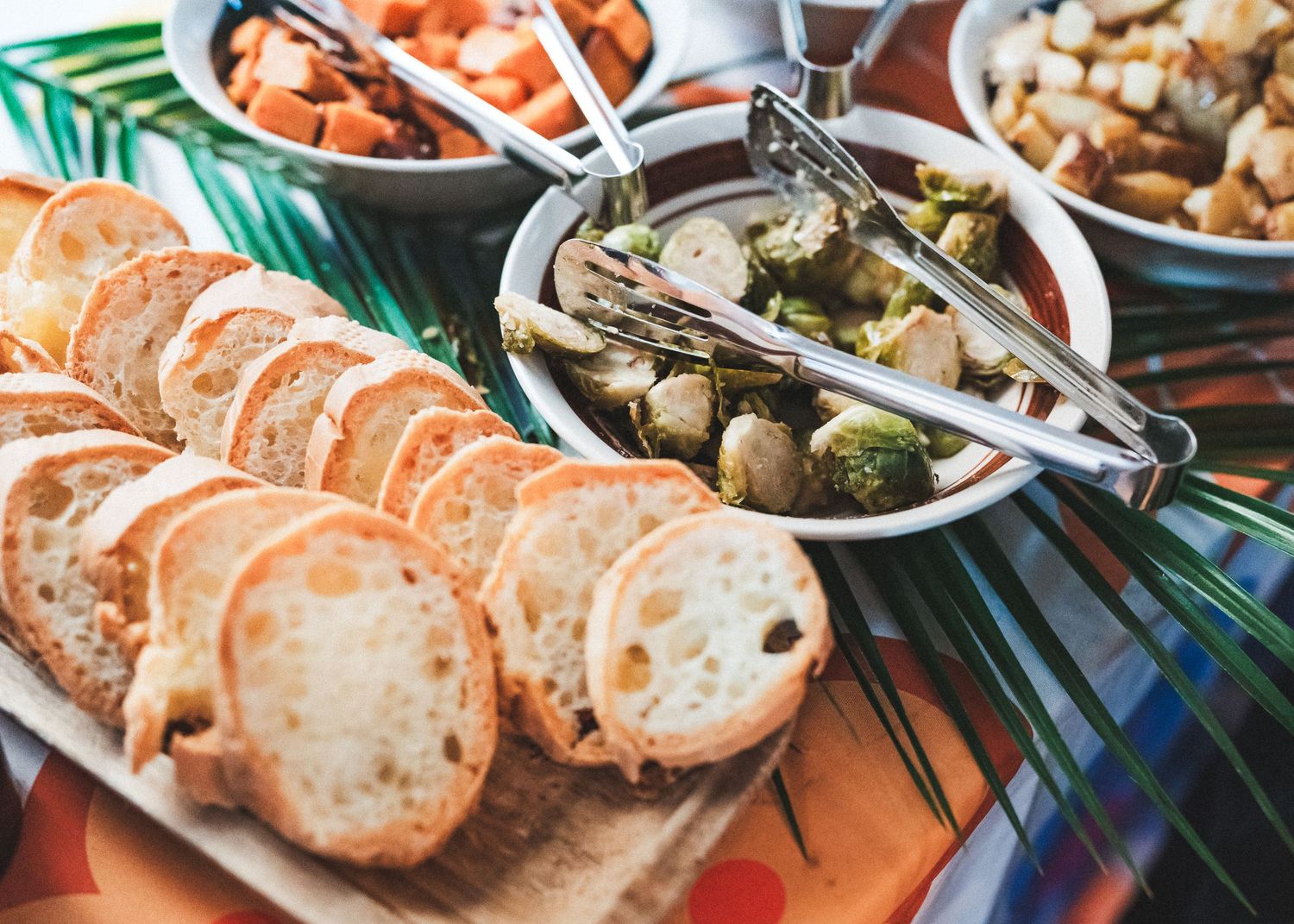 Fresh baguette slices served on wooden board with olives and various appetizers on palm leaf decoration.