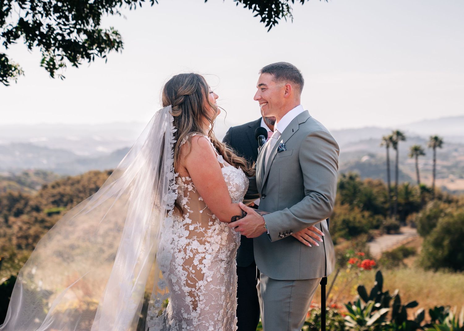 A romantic wedding couple embraces on a hilltop overlooking a scenic mountain landscape with palm trees in the distance.