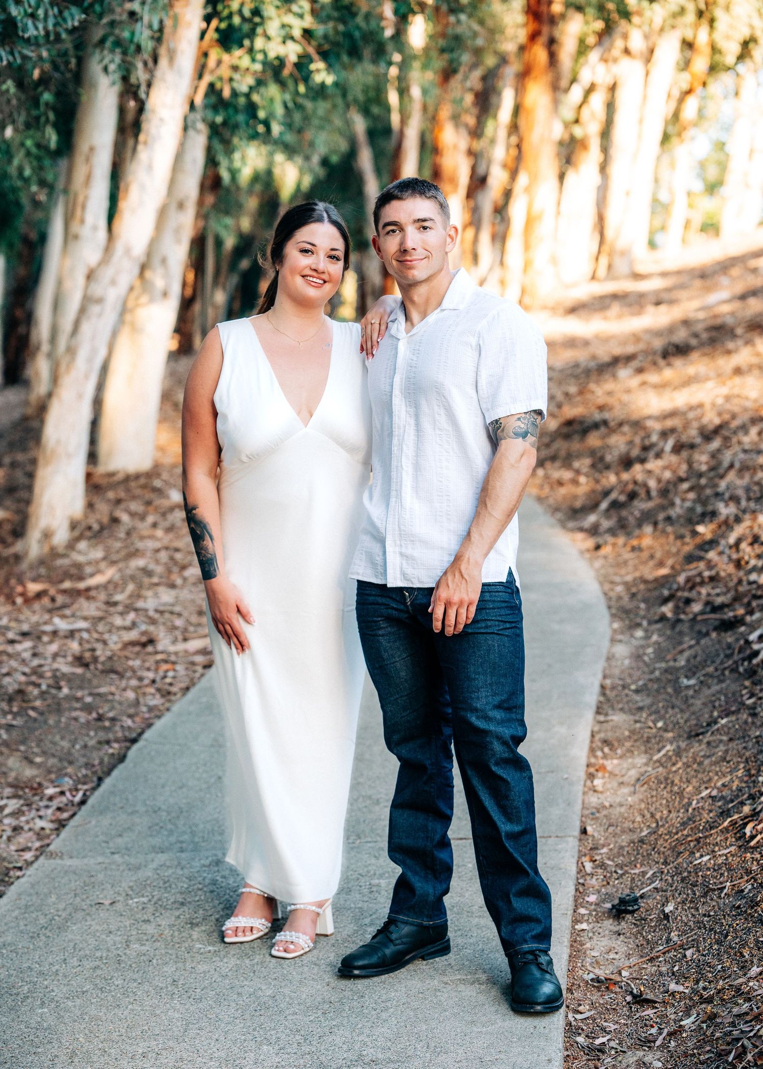 A couple poses on a wooded path together, with sunlight filtering through trees behind them.