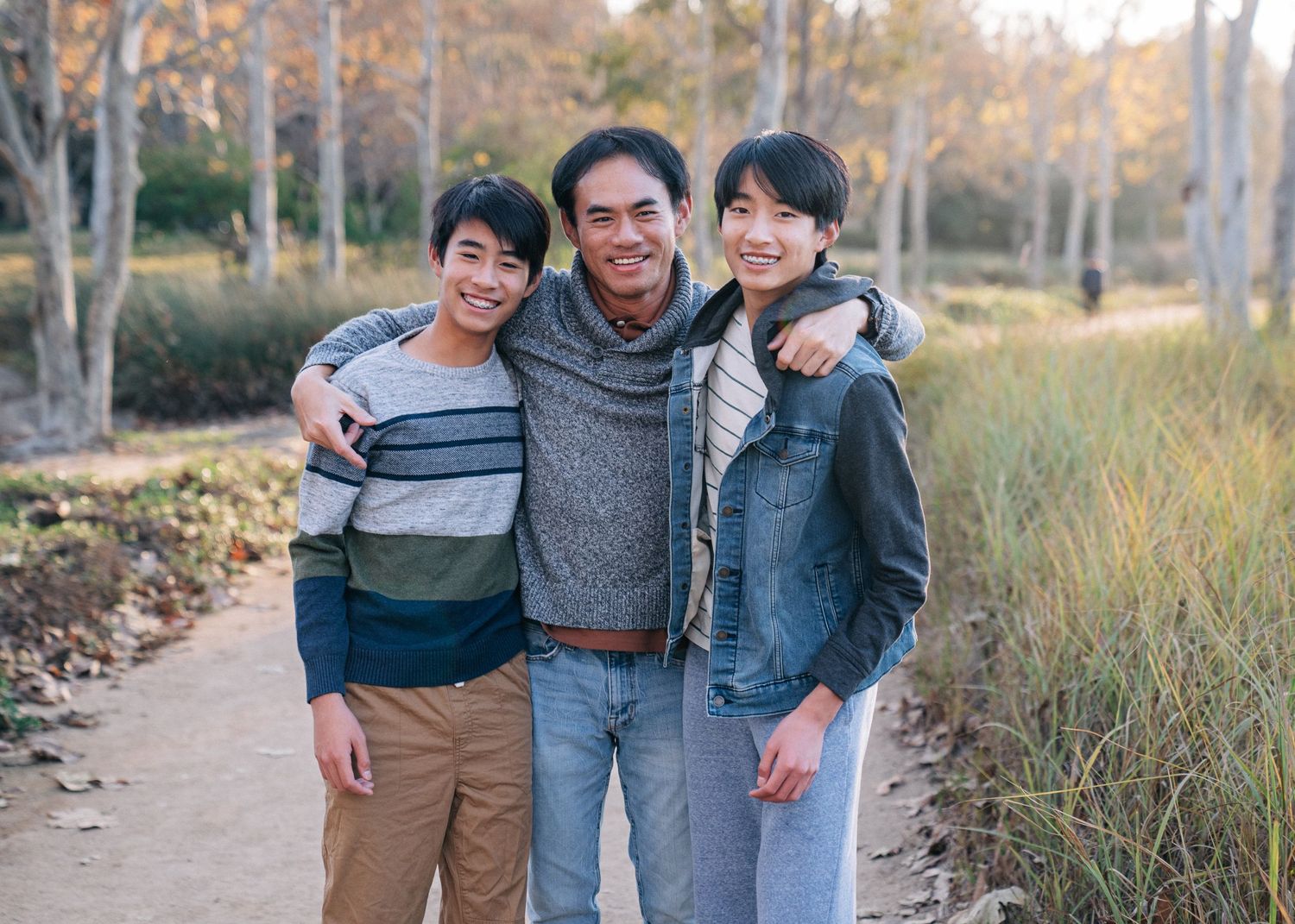 Three people wearing casual clothing pose together on a hiking trail surrounded by autumn trees and tall grass.