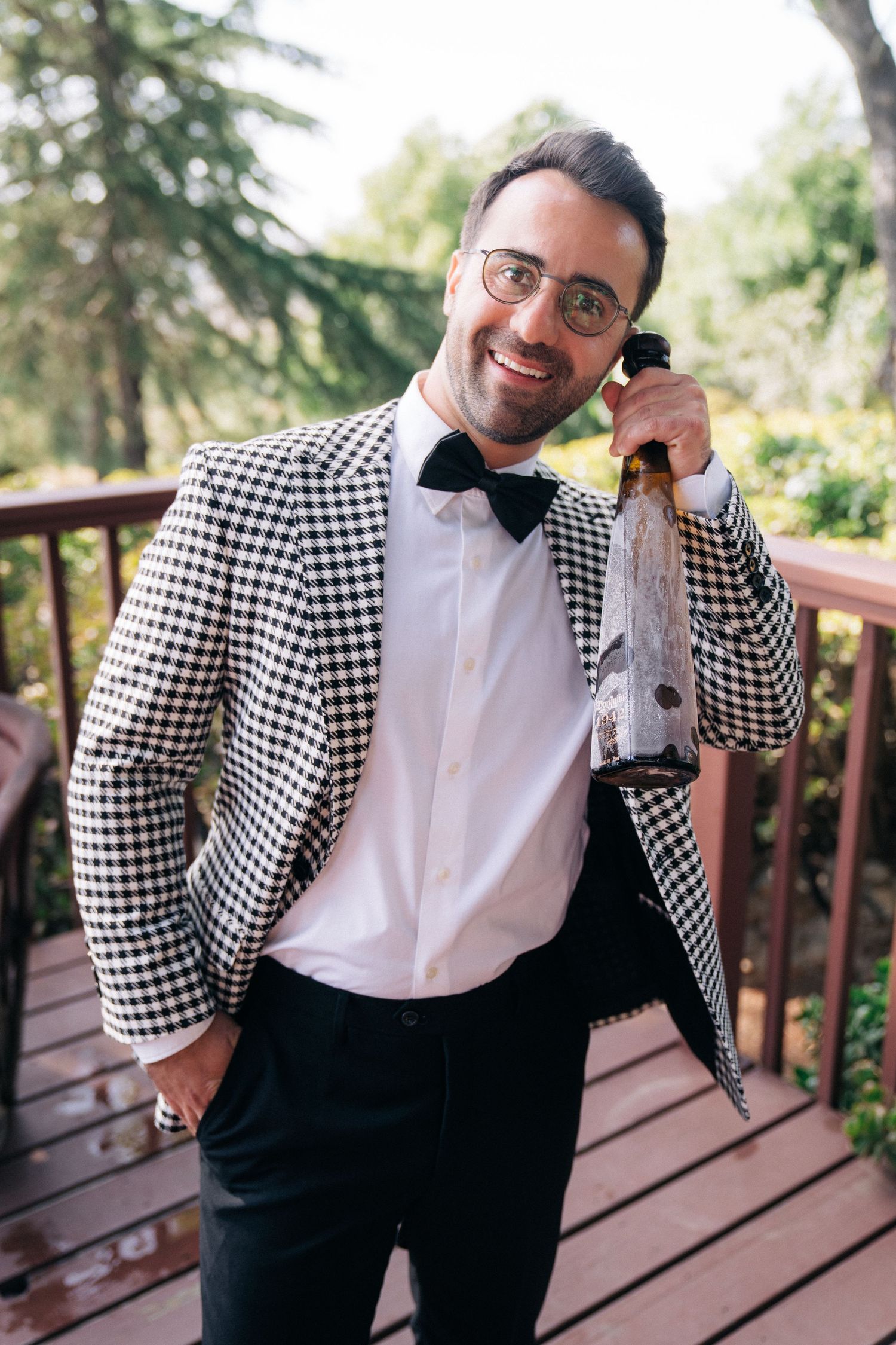 Groom in checkered suit jacket and bow tie poses on wooden deck.