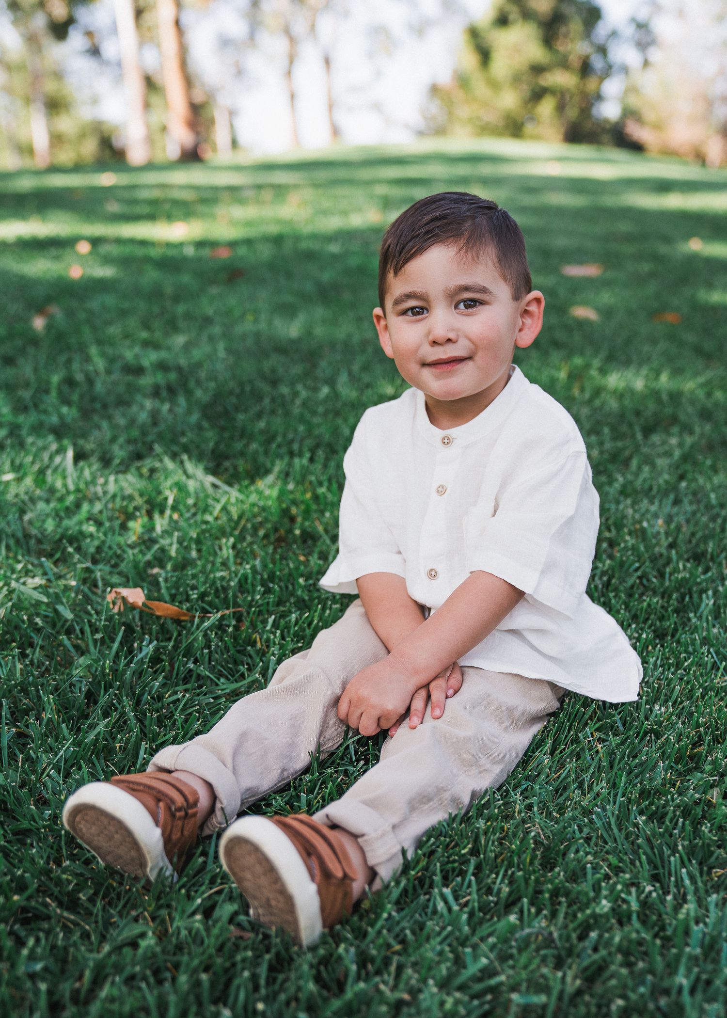 Young child sits on grass in a white shirt and beige pants with brown shoes in an outdoor park setting.