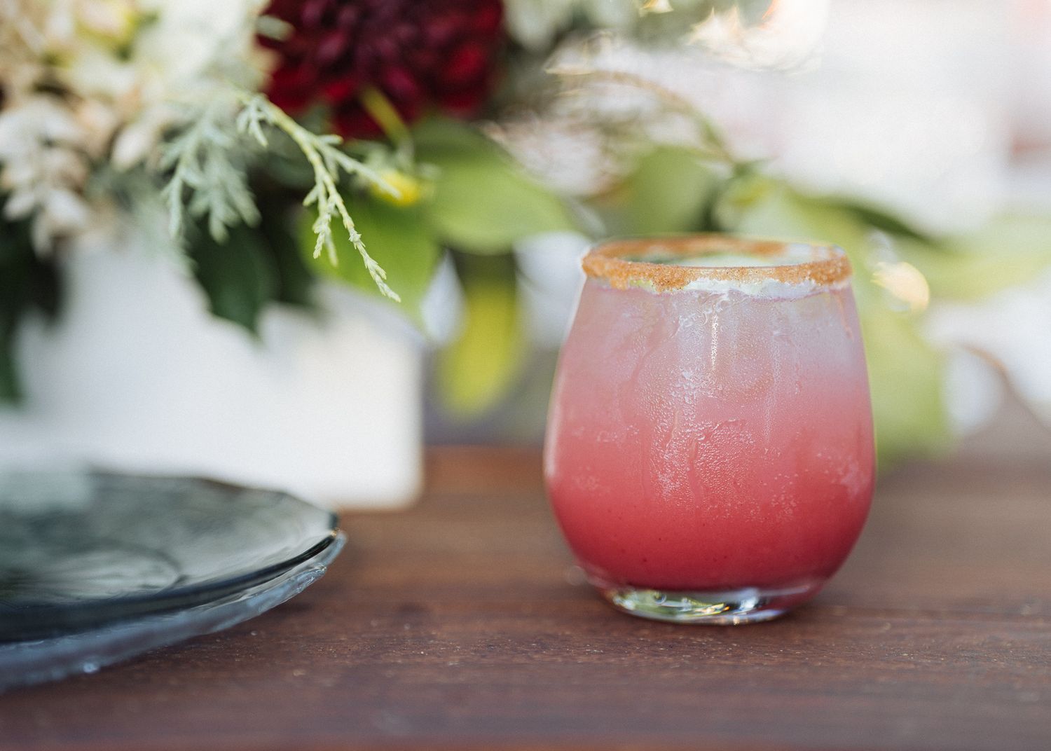 A pink cocktail served in a curved glass with floral decorations on a wooden table.
