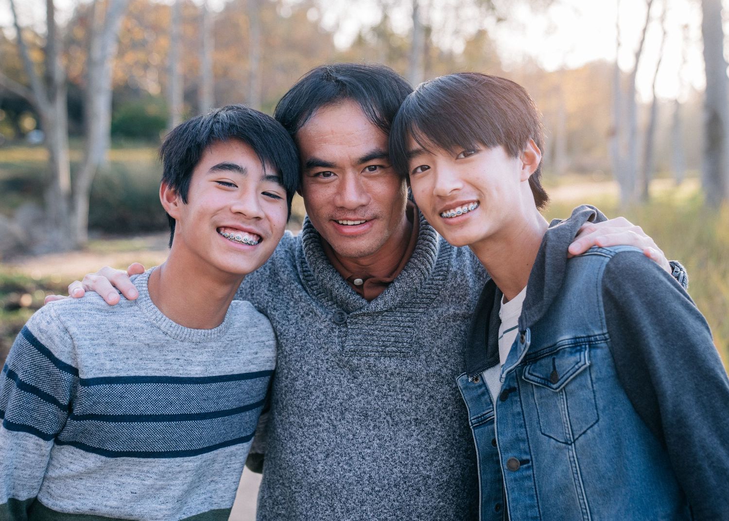 Three people wearing casual clothing smile together outdoors during autumn with trees in the background.