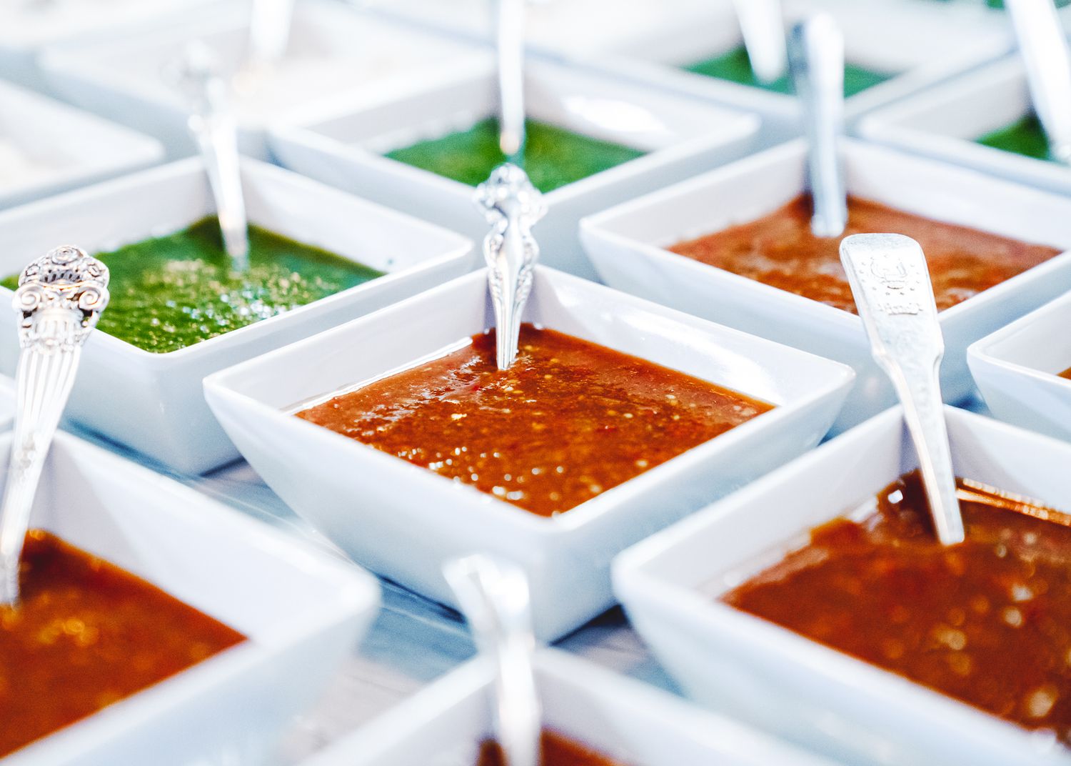 Various colorful dipping sauces and condiments served in small white square dishes with plastic spoons.