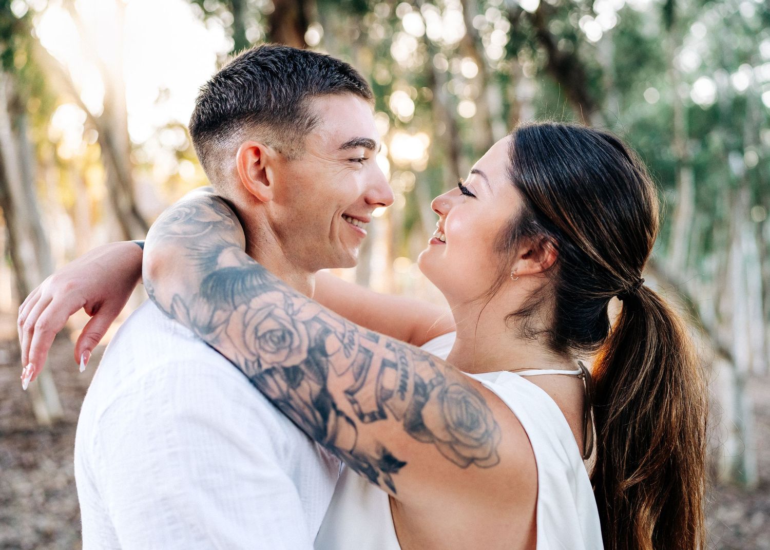 A loving couple embraces outdoors at sunset, with visible tattoos on his arm as they share an affectionate moment.
