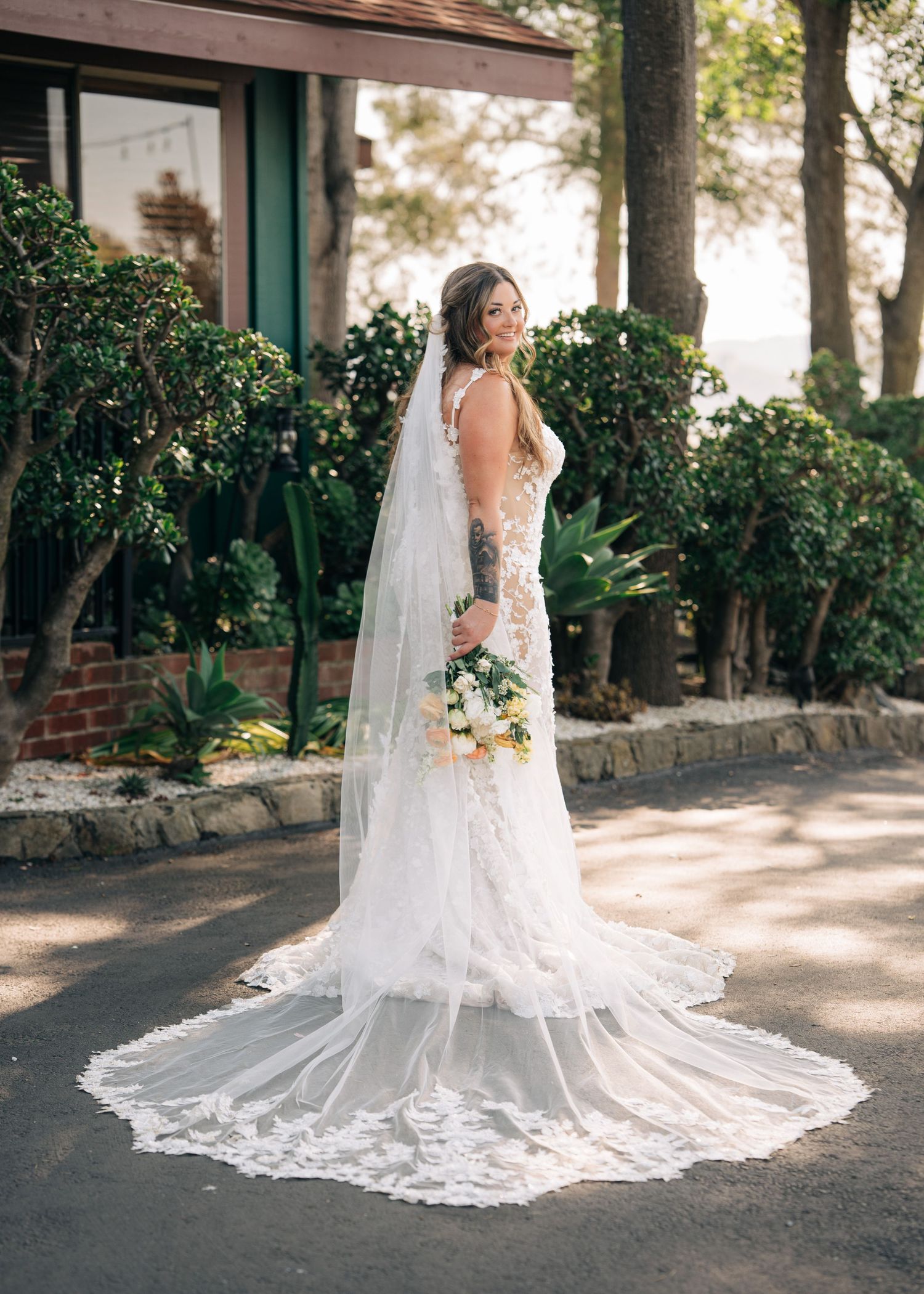 Beautiful bride in lace wedding dress with cathedral length veil poses outdoors against greenery and building exterior.