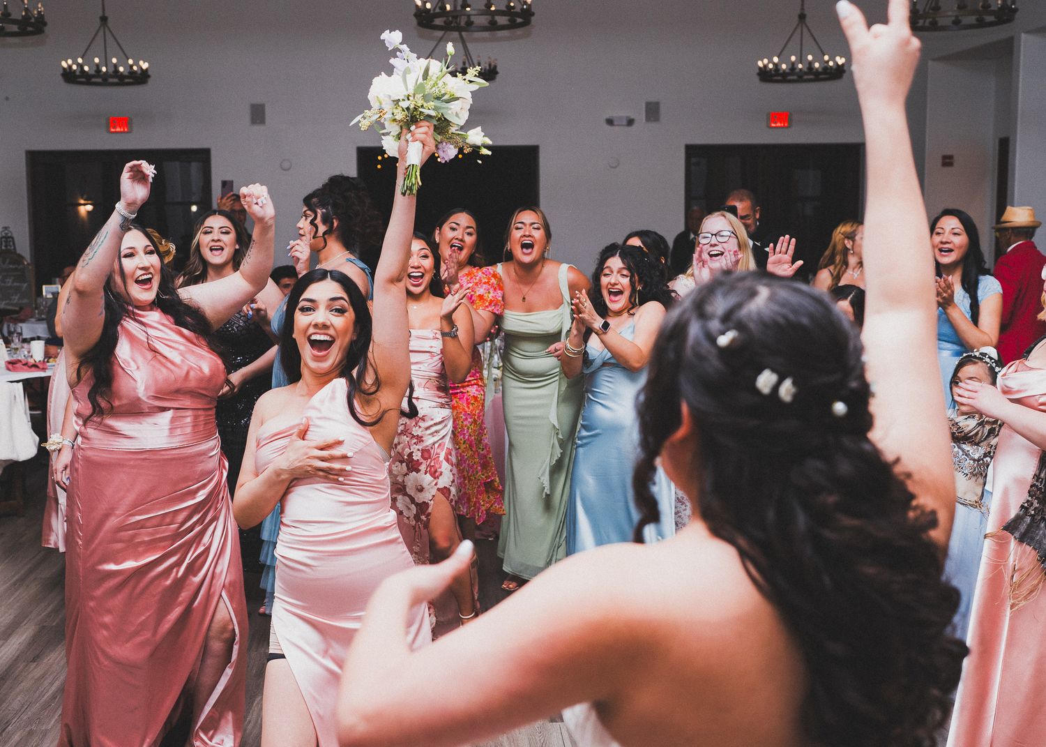 Wedding reception guests in pink and pastel dresses celebrate joyfully on the dance floor with raised arms and bouquet.