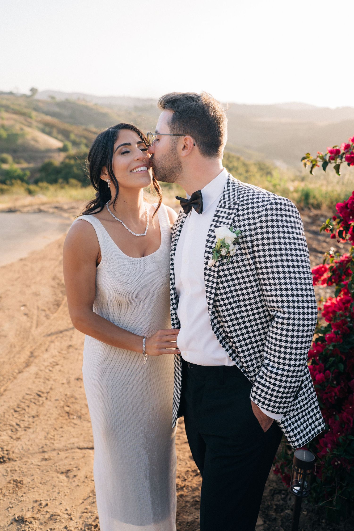 A romantic wedding photo captured on a hilltop dirt road with beautiful red flowers at sunset.