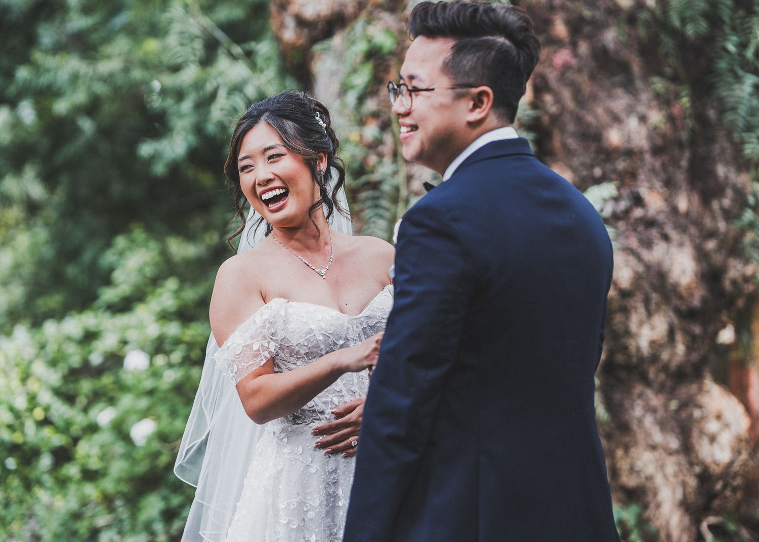 Wedding couple and bridal party laughing and celebrating in a garden setting against greenery.
