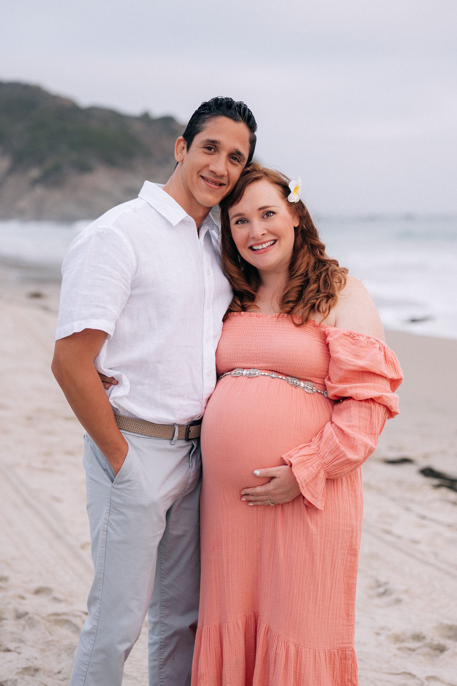 A couple in white and coral attire shares tender moments during a beach maternity photoshoot at sunset.