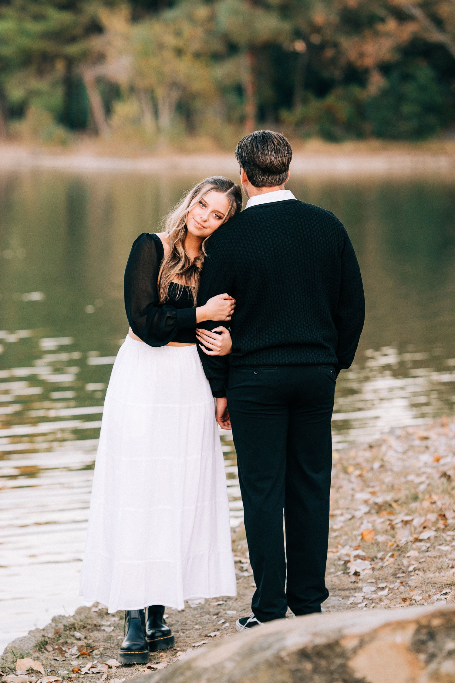 A couple embraces beside a serene lake with autumn foliage in the background.