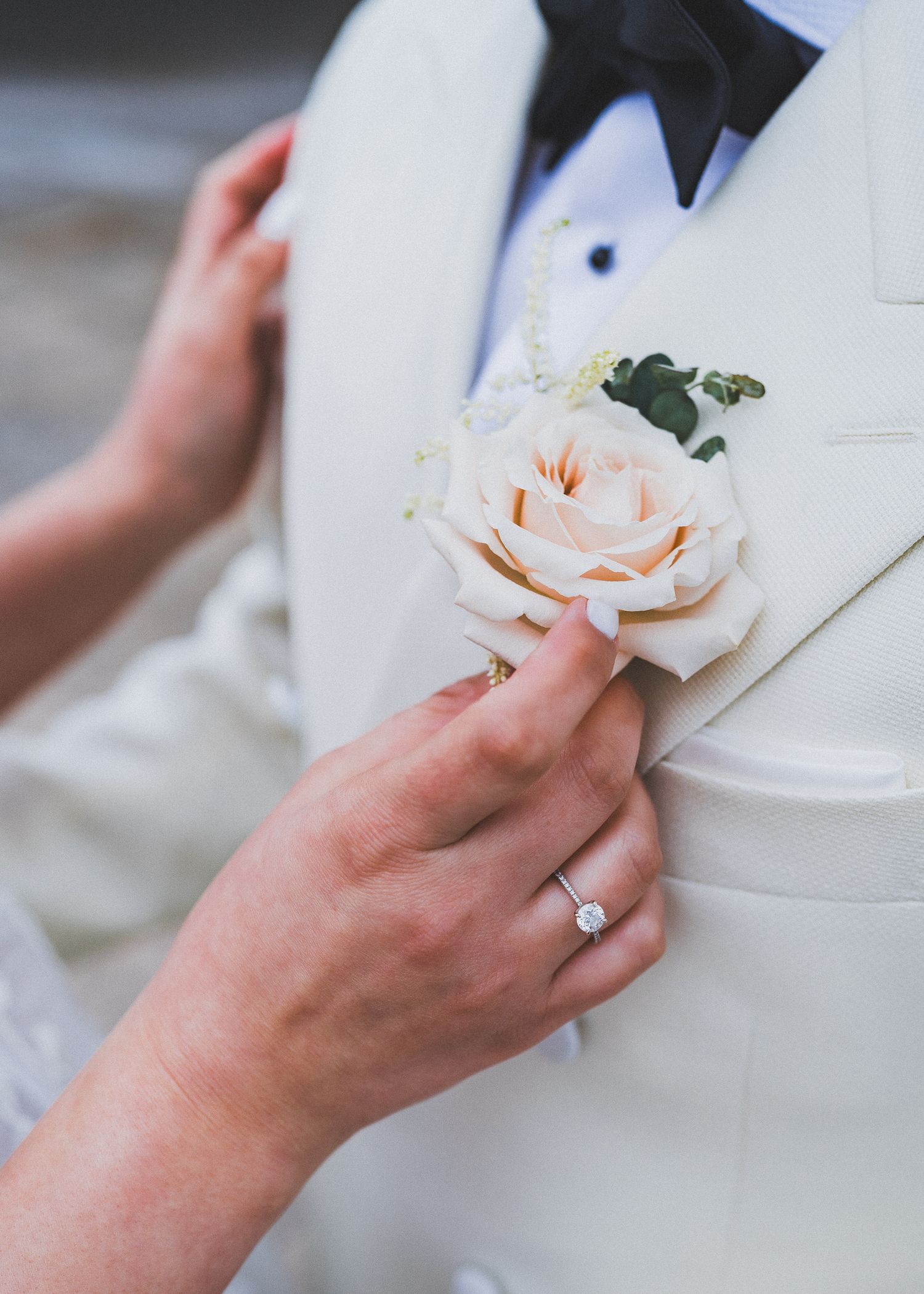 Someone wearing an engagement ring arranges a delicate peach-colored rose boutonniere against a white formal garment.