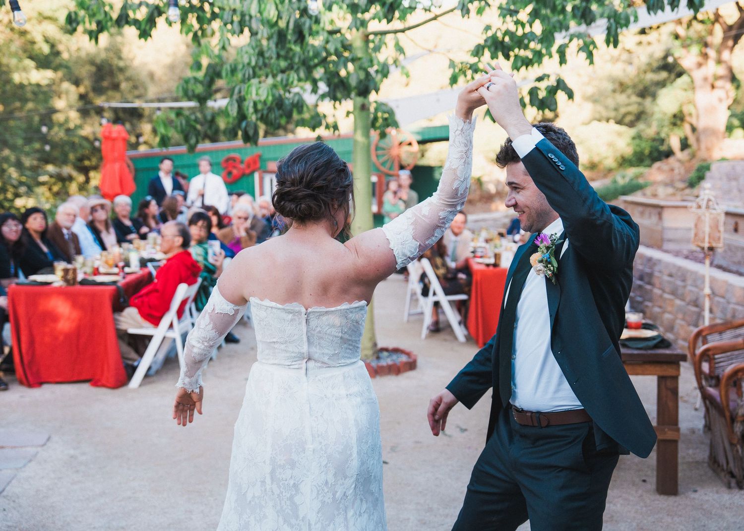 A joyful couple dances at their outdoor wedding reception with red table decor visible in the background.