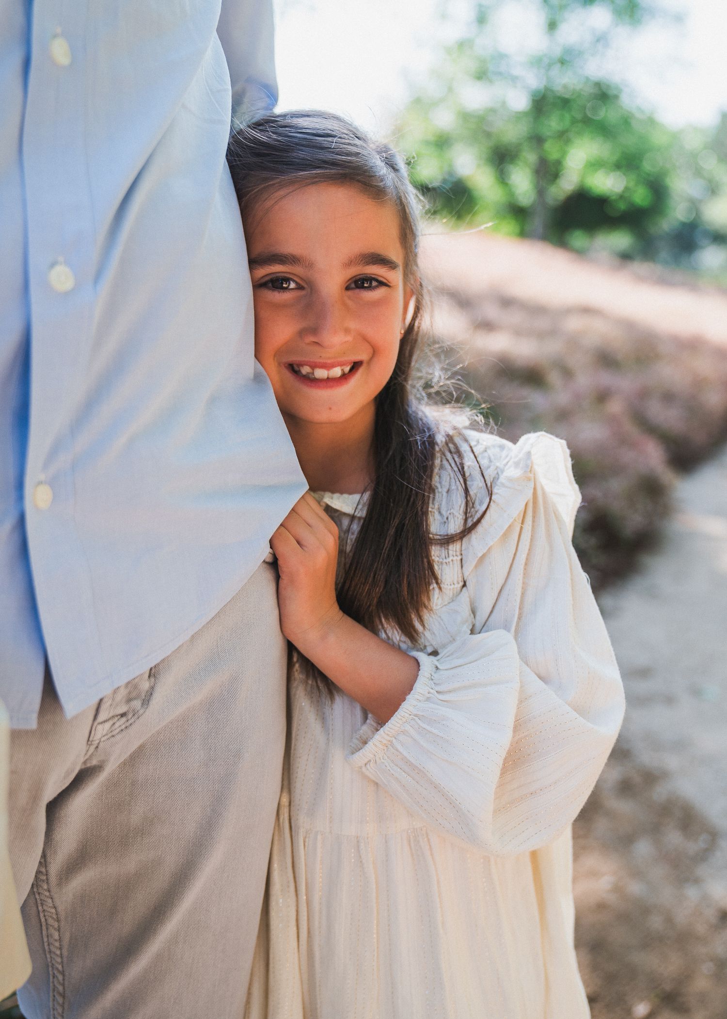 A young person in a white dress peeks out playfully from behind someone wearing a white shirt outdoors.
