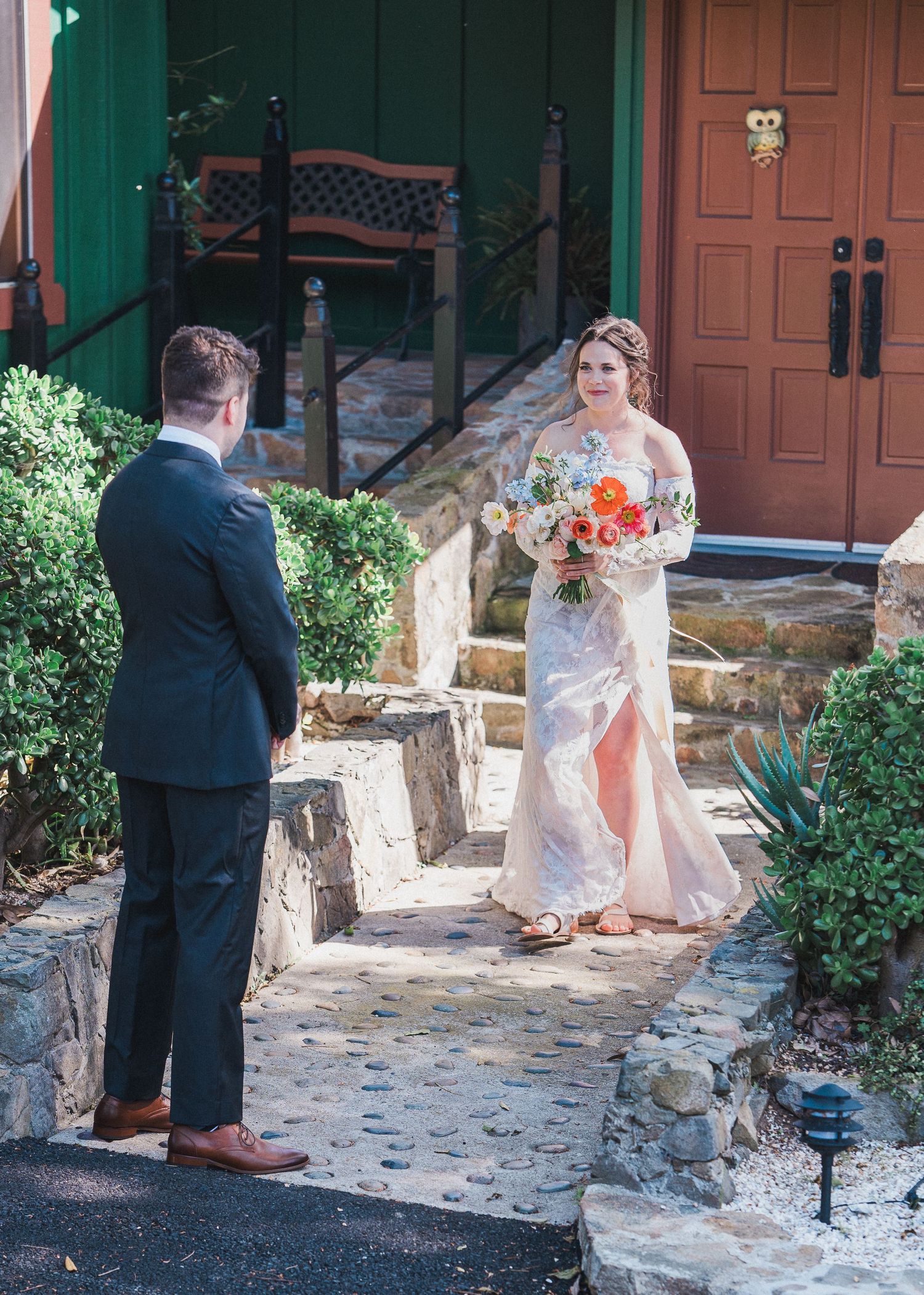 Outdoor wedding scene with a couple on stone steps by a rustic building with orange floral bouquet.