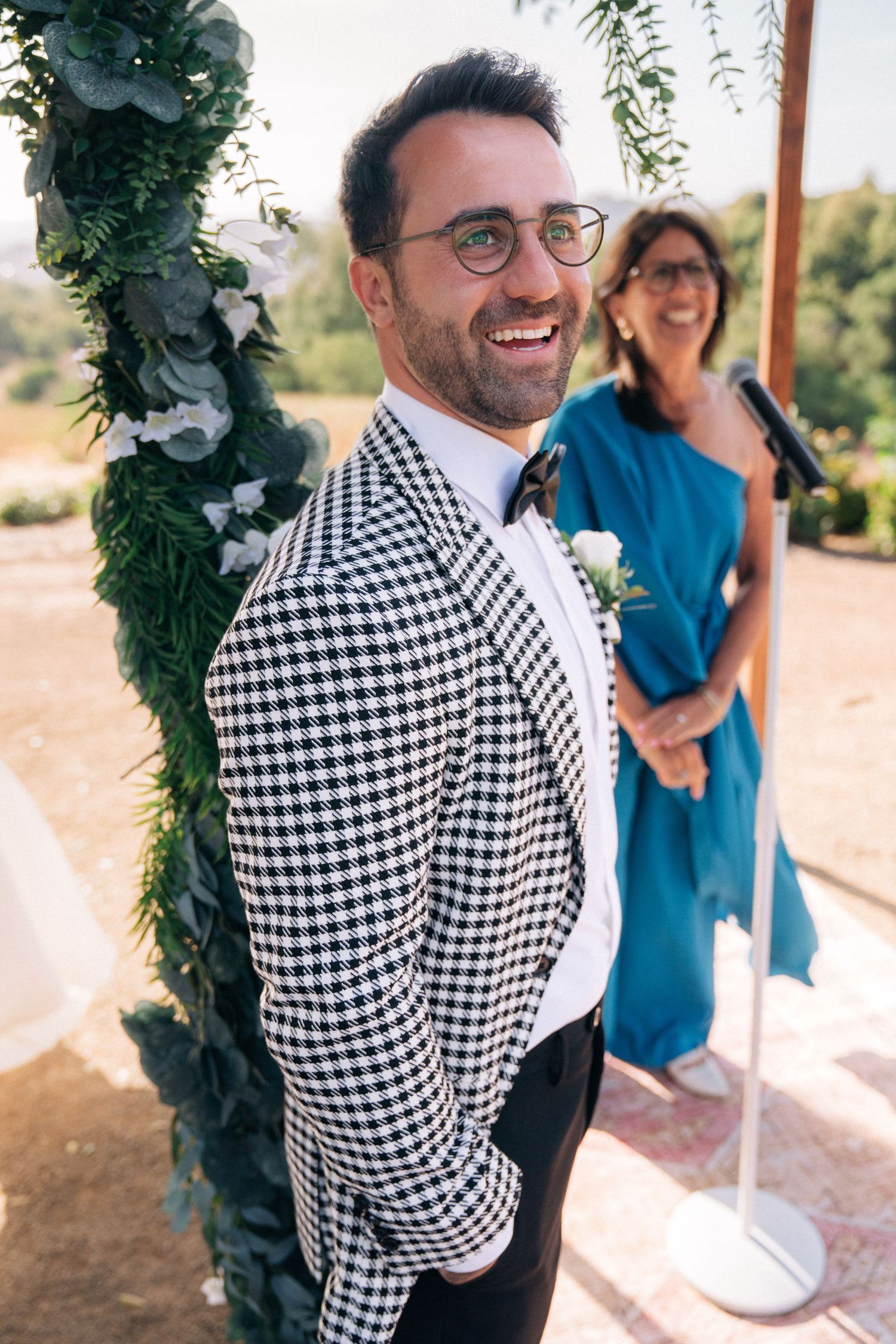 Groom wearing distinctive black and white checkered suit jacket smiles during outdoor ceremony.