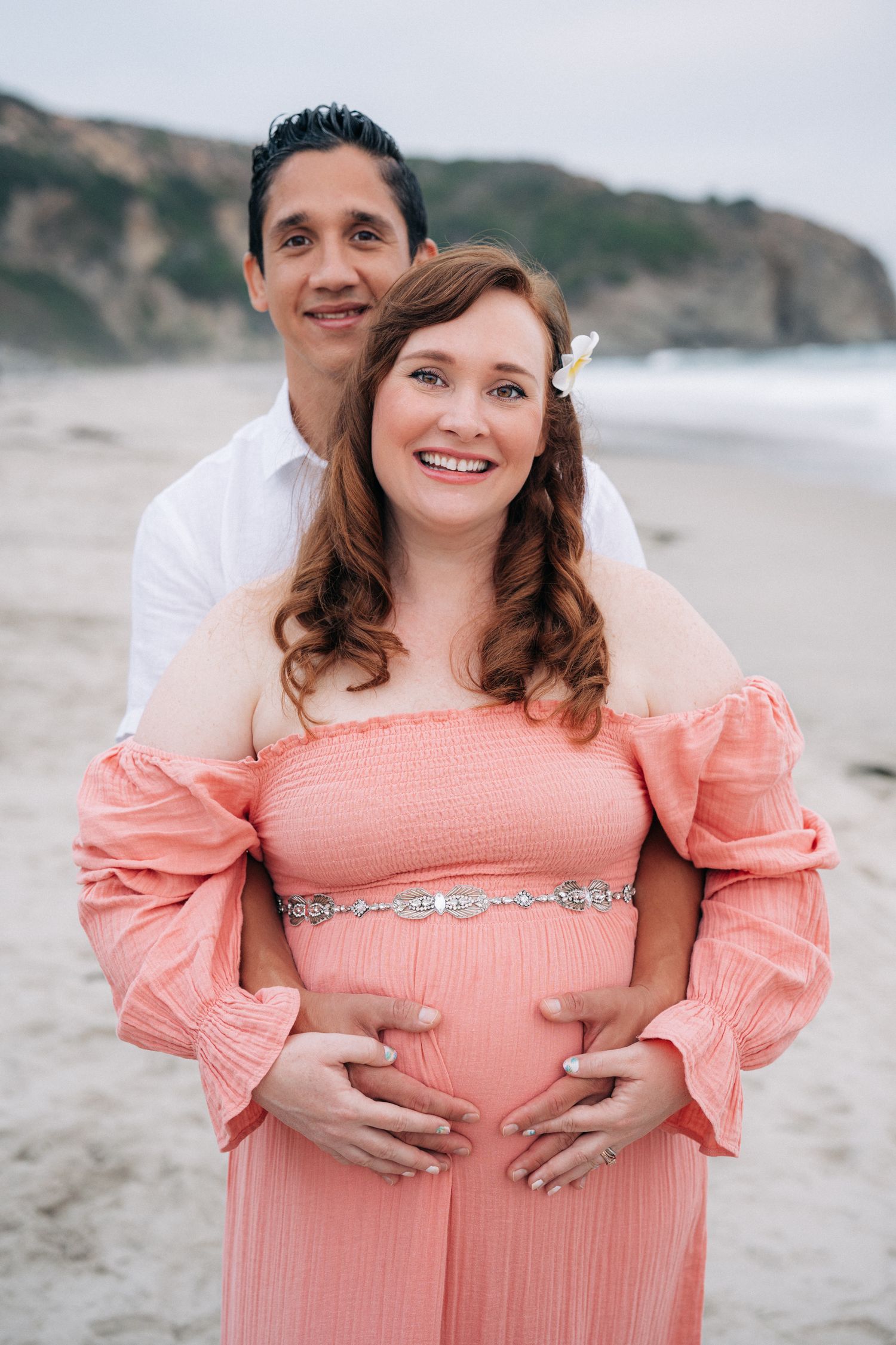 A couple wearing white and peach outfits poses lovingly for maternity photos on a cloudy beach.