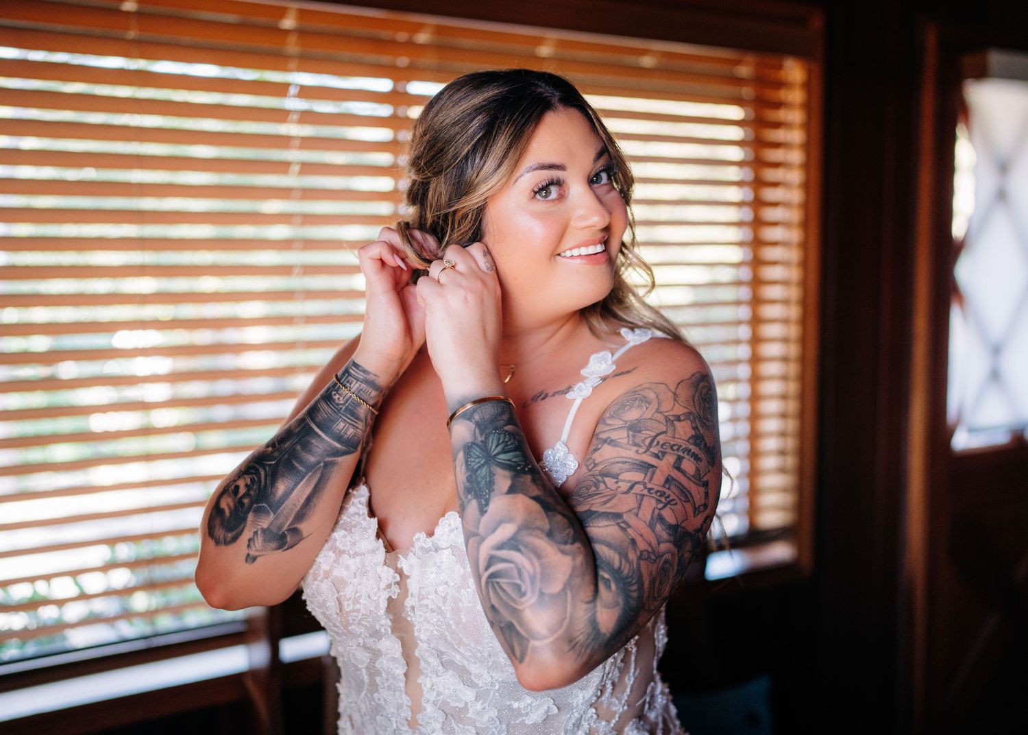 A person in a lace wedding dress adjusts their earrings near a window with wooden blinds.