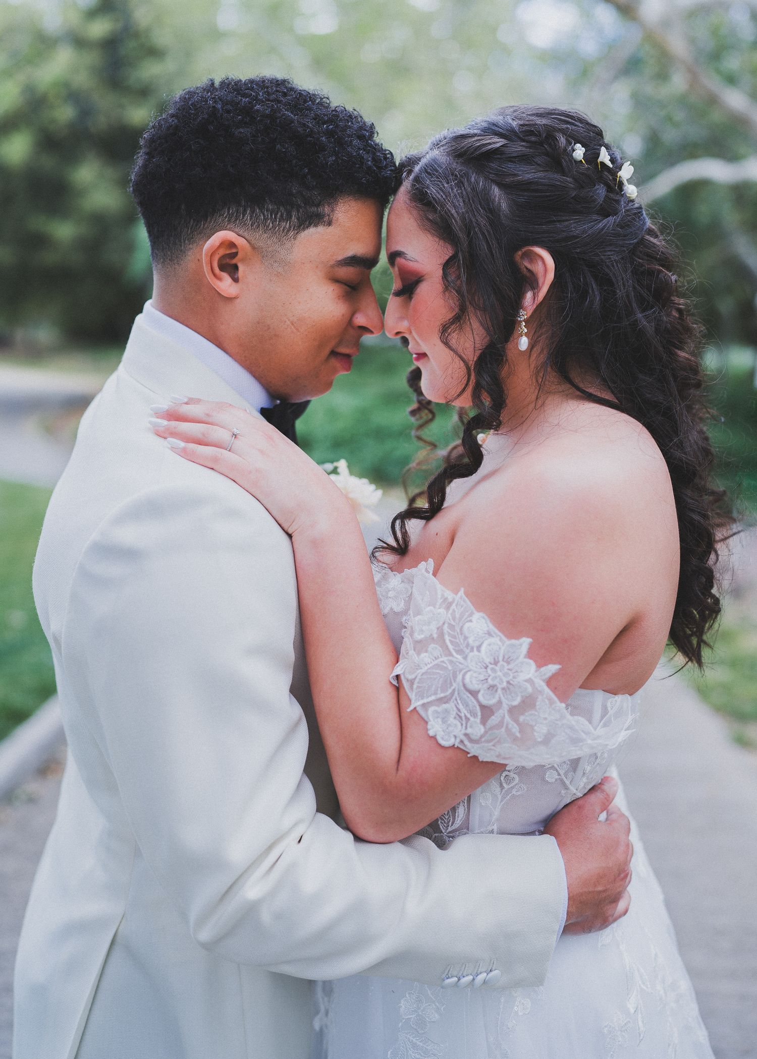A romantic wedding photo showing a tender moment between newlyweds in white attire against a soft green outdoor background.