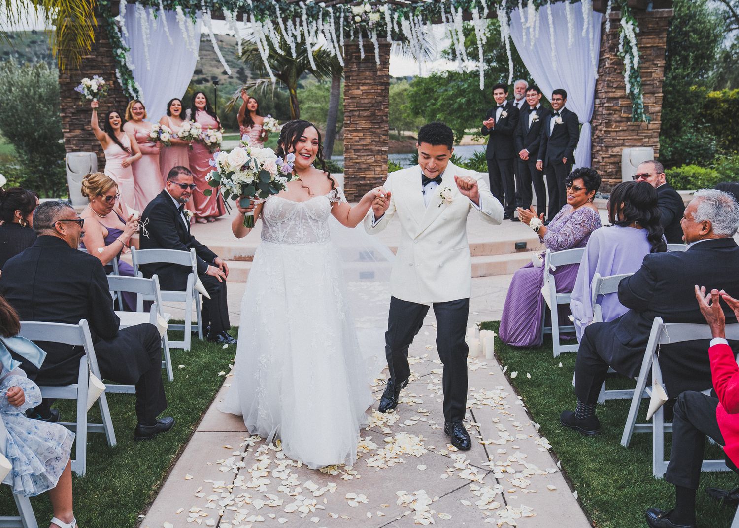 A joyful wedding ceremony moment as newlyweds dance down a rose petal covered aisle lined with white chairs and palm trees.