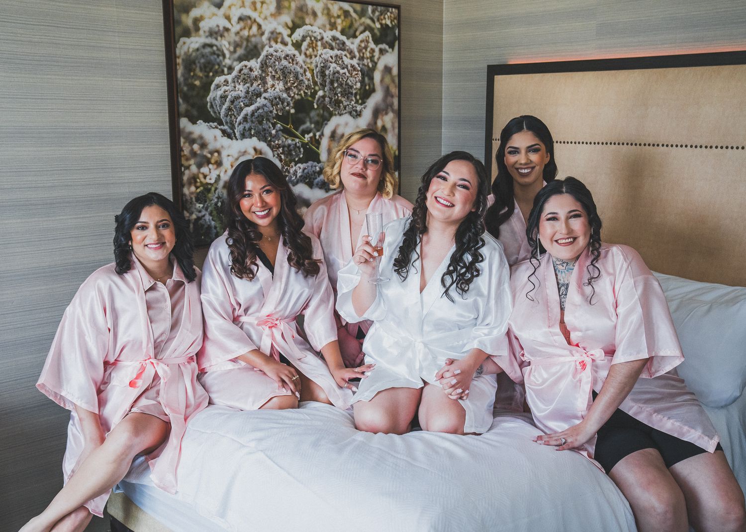 A group wearing matching pink robes sits together on a hotel bed smiling at the camera.