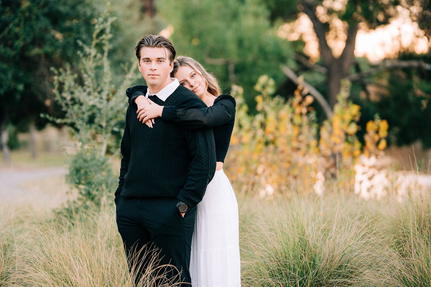 A couple embracing in front of tall grass during sunset wearing black and white outfits.