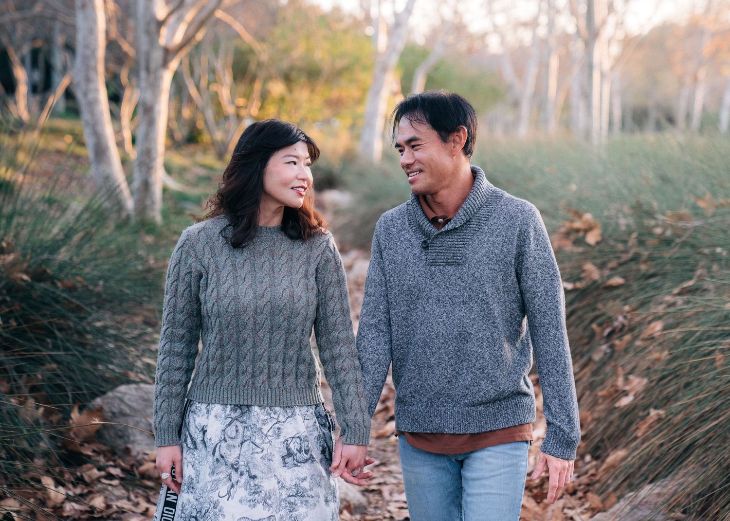 Two people in gray sweaters walk together while laughing on an autumn nature trail surrounded by dried grass and trees.