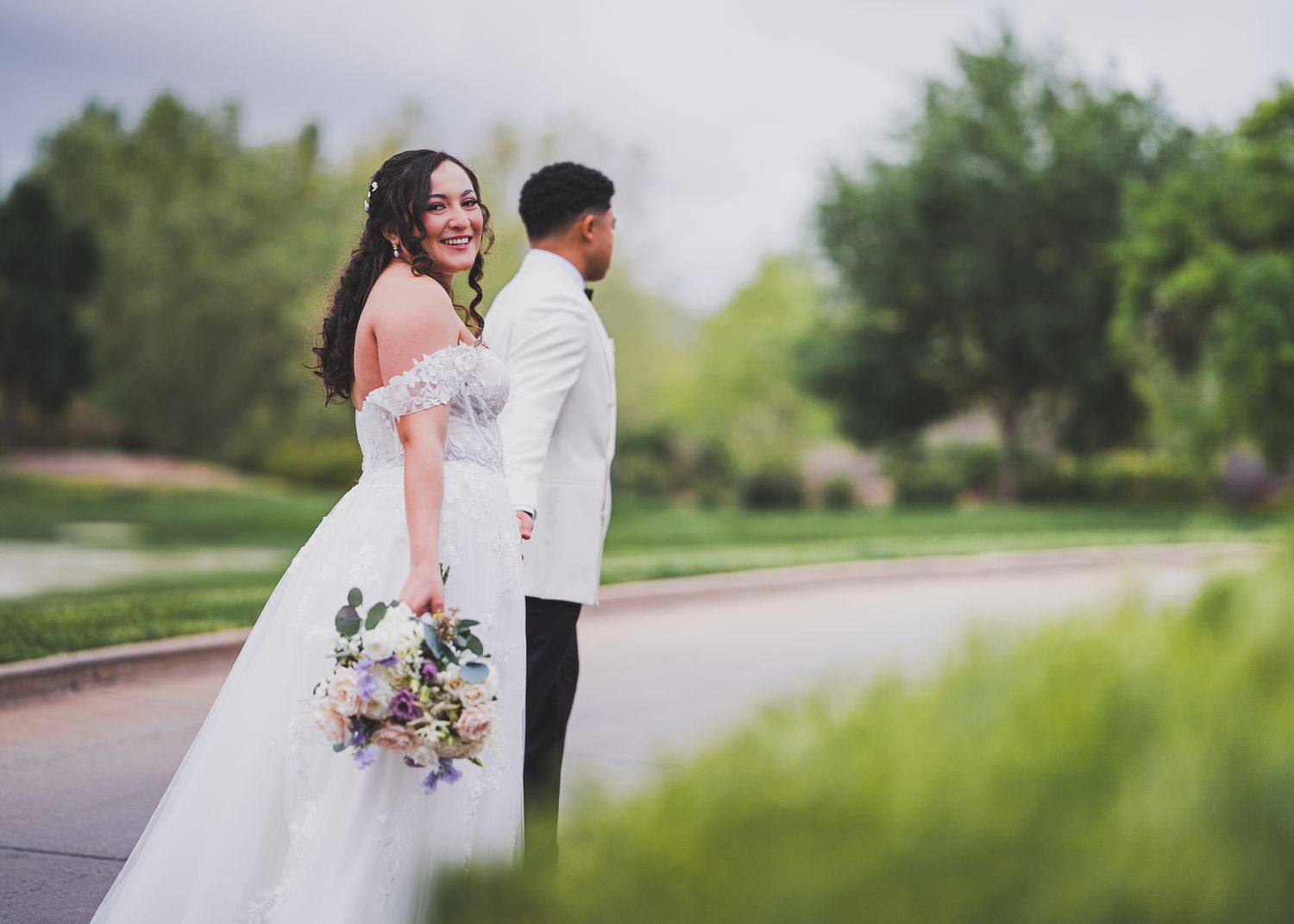 A couple in wedding attire share a joyful moment together on a tree-lined path while holding a purple floral bouquet.