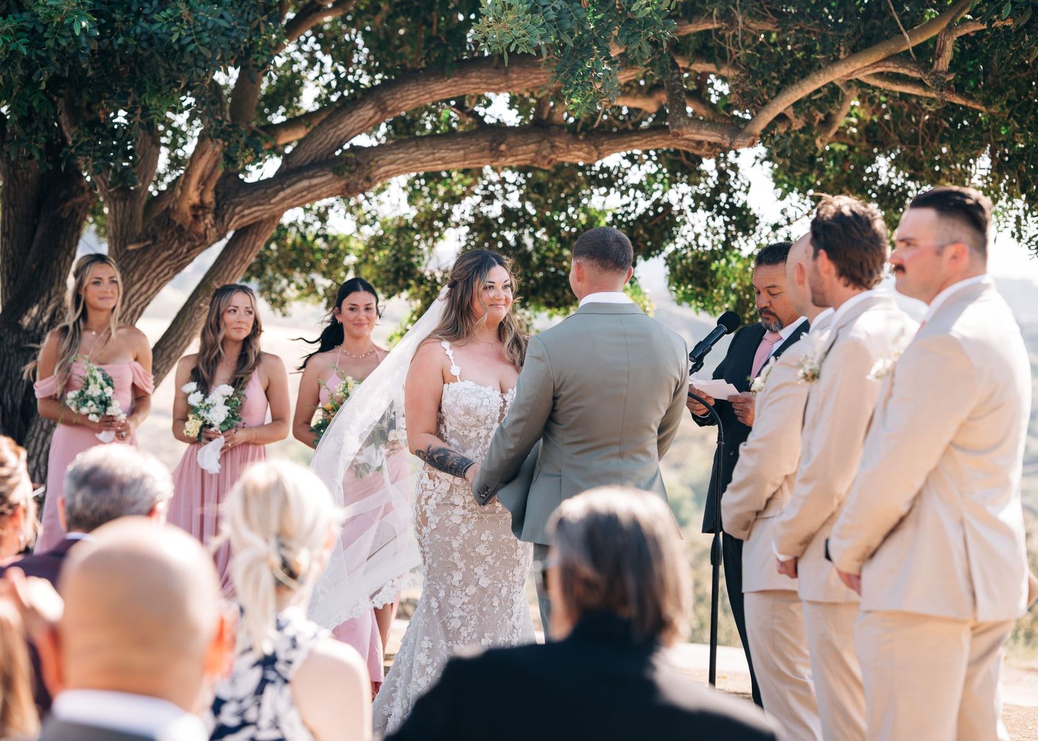 An outdoor wedding ceremony under a large tree with the bridal party gathered in beige and pink attire during a sunny day.