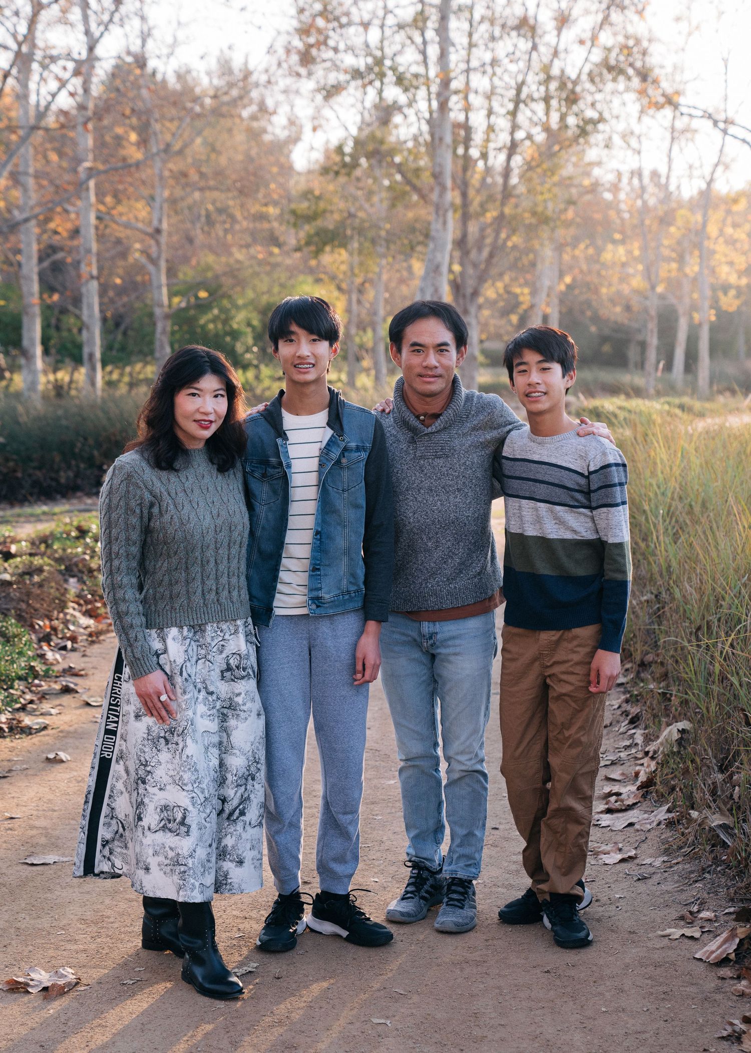 A small group in casual autumn attire poses together on a dirt path surrounded by fall foliage and trees.