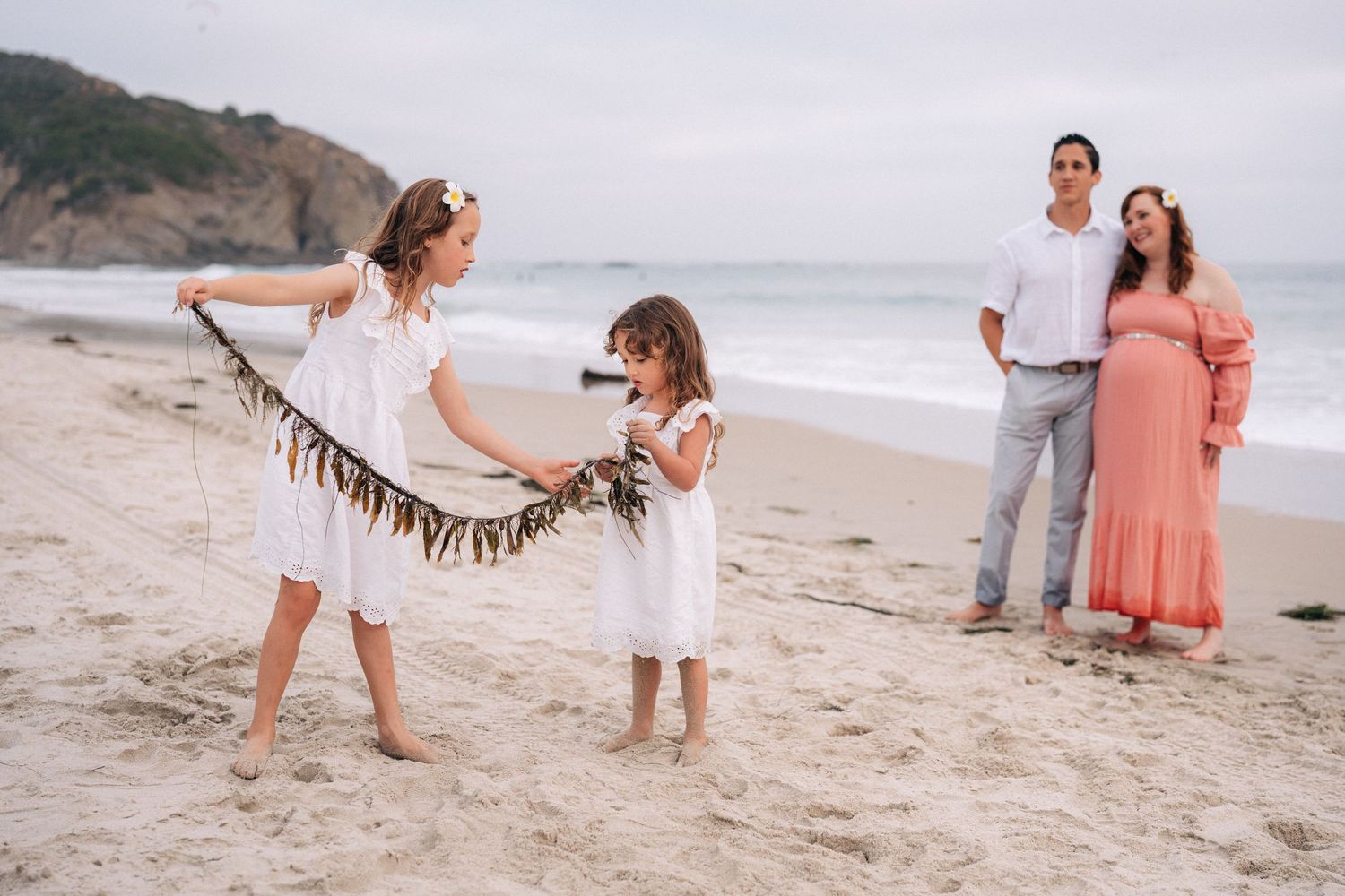 Casual beach photoshoot featuring family members in white and coral outfits near the ocean.