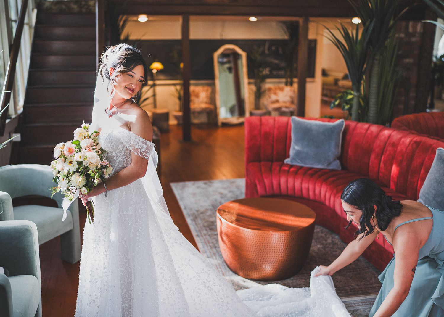 Bride with bouquet poses near red velvet couch in elegant indoor setting.
