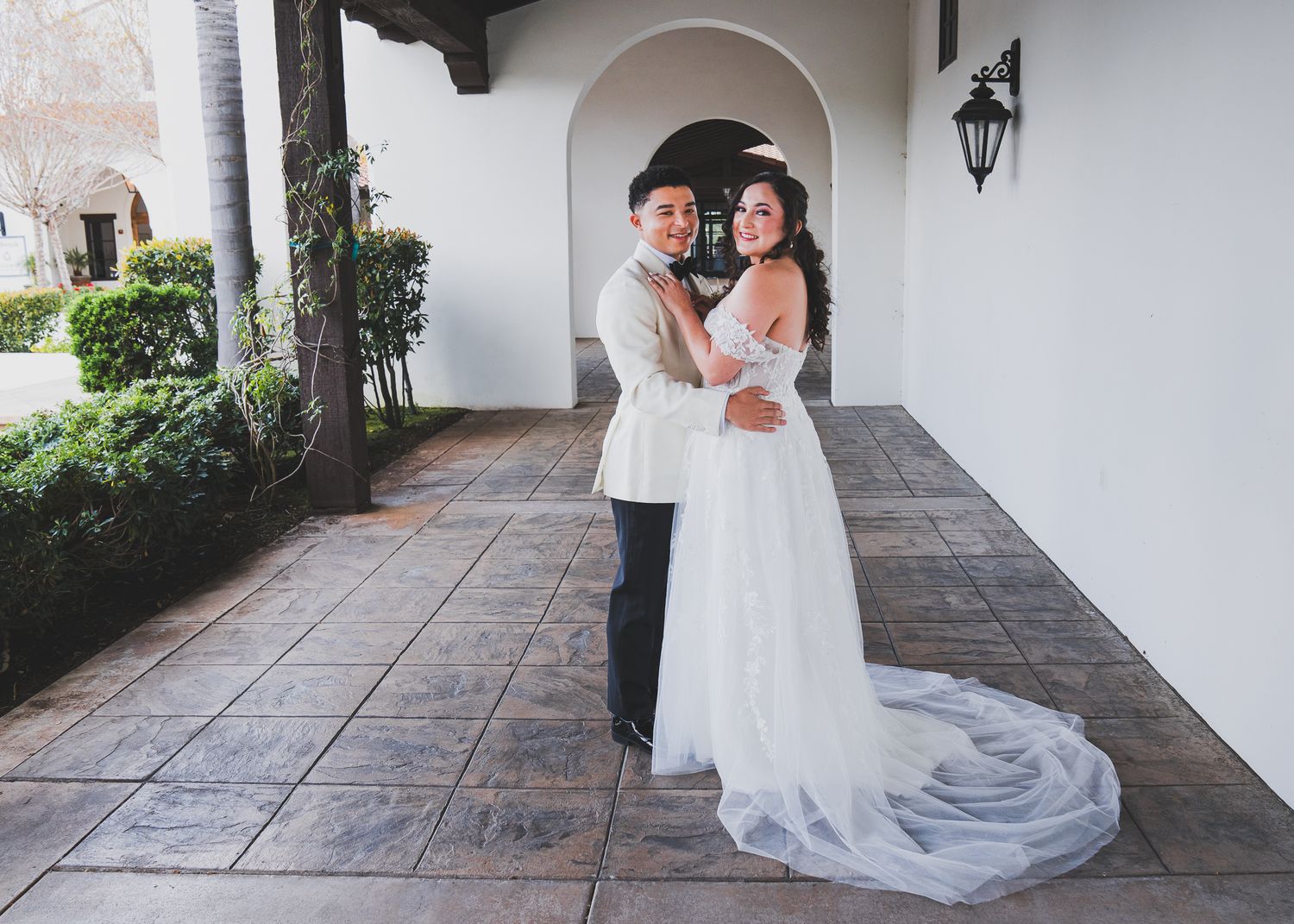 A couple poses together at an elegant venue with white stucco walls and archways, the bride wearing a flowing gown with train.