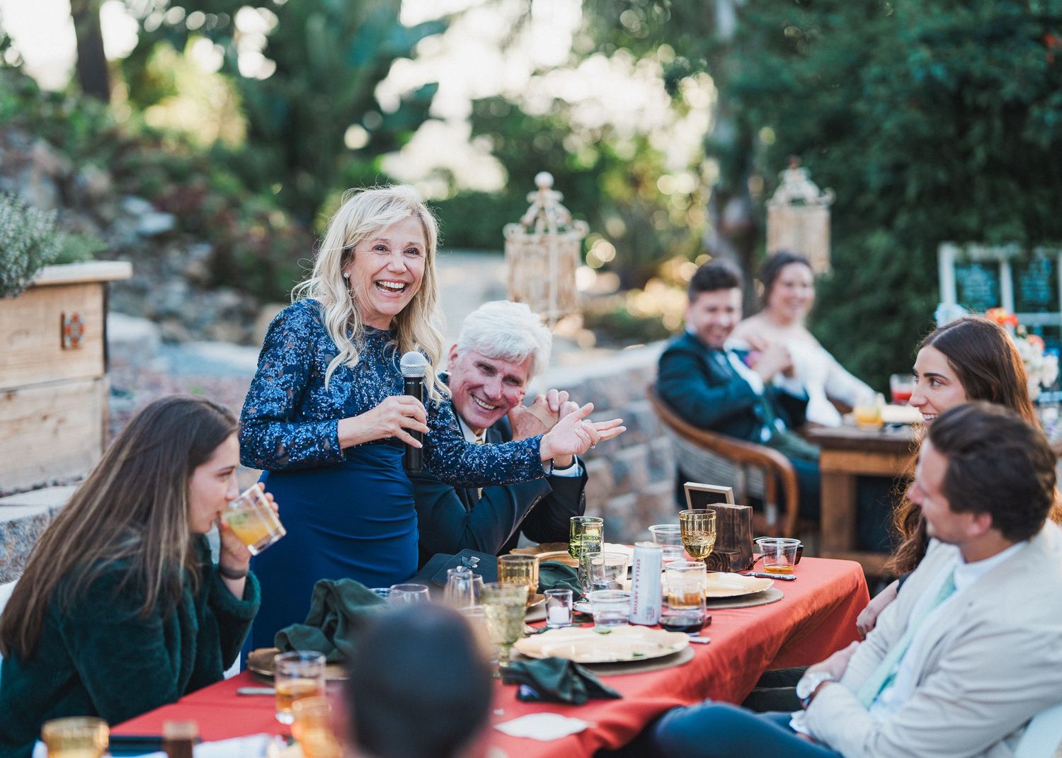 Wedding guests enjoy a casual outdoor reception dinner at rustic wooden tables.