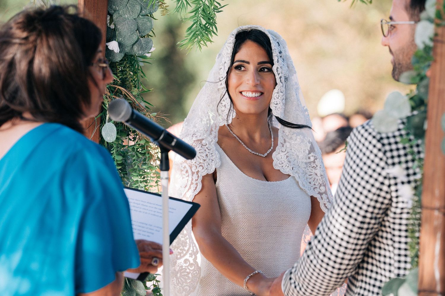 Wedding celebration scene with decorative greenery and guests in bright outdoor setting.