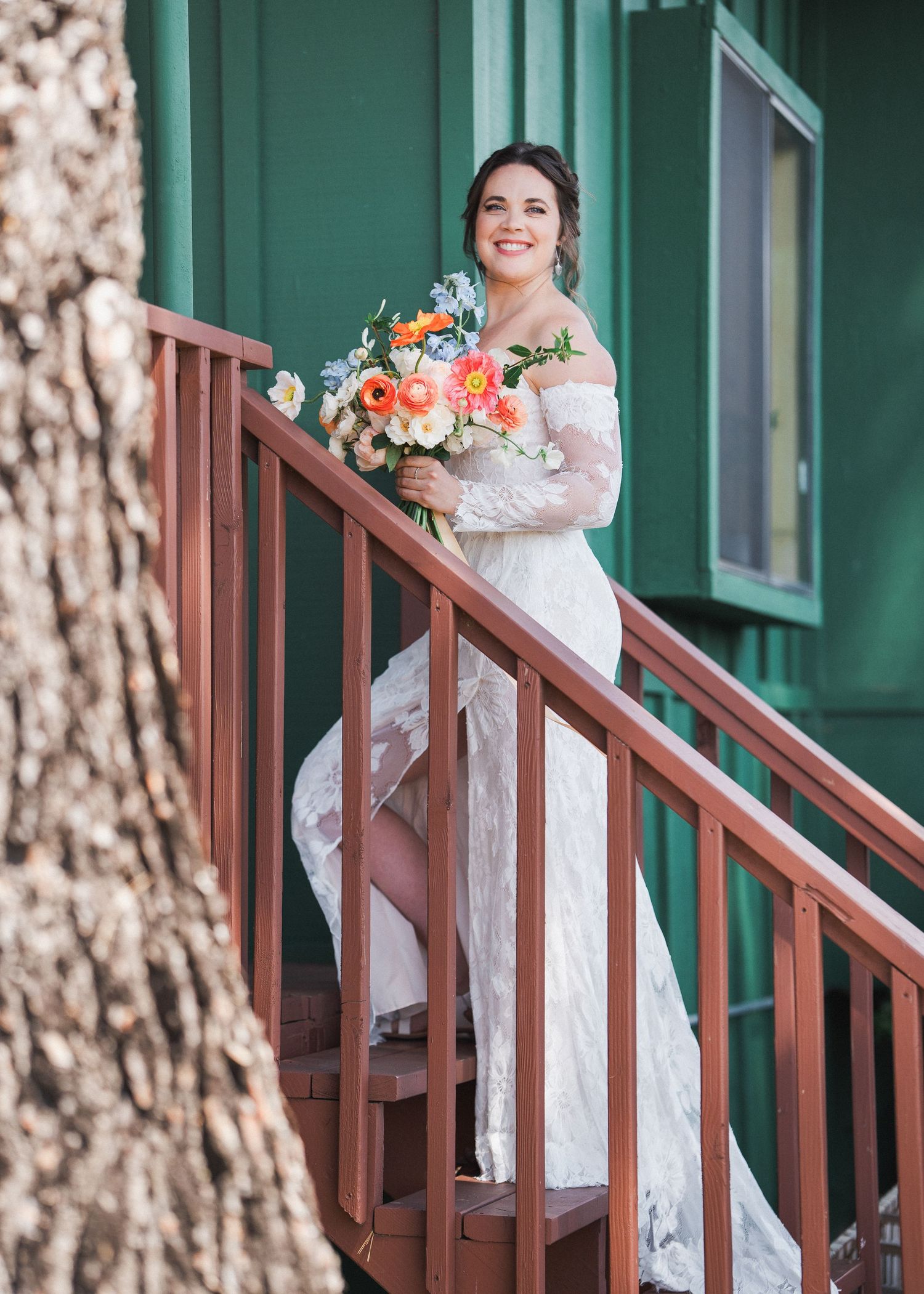 Bride in off-shoulder lace dress stands on green wooden staircase holding coral and white bouquet.
