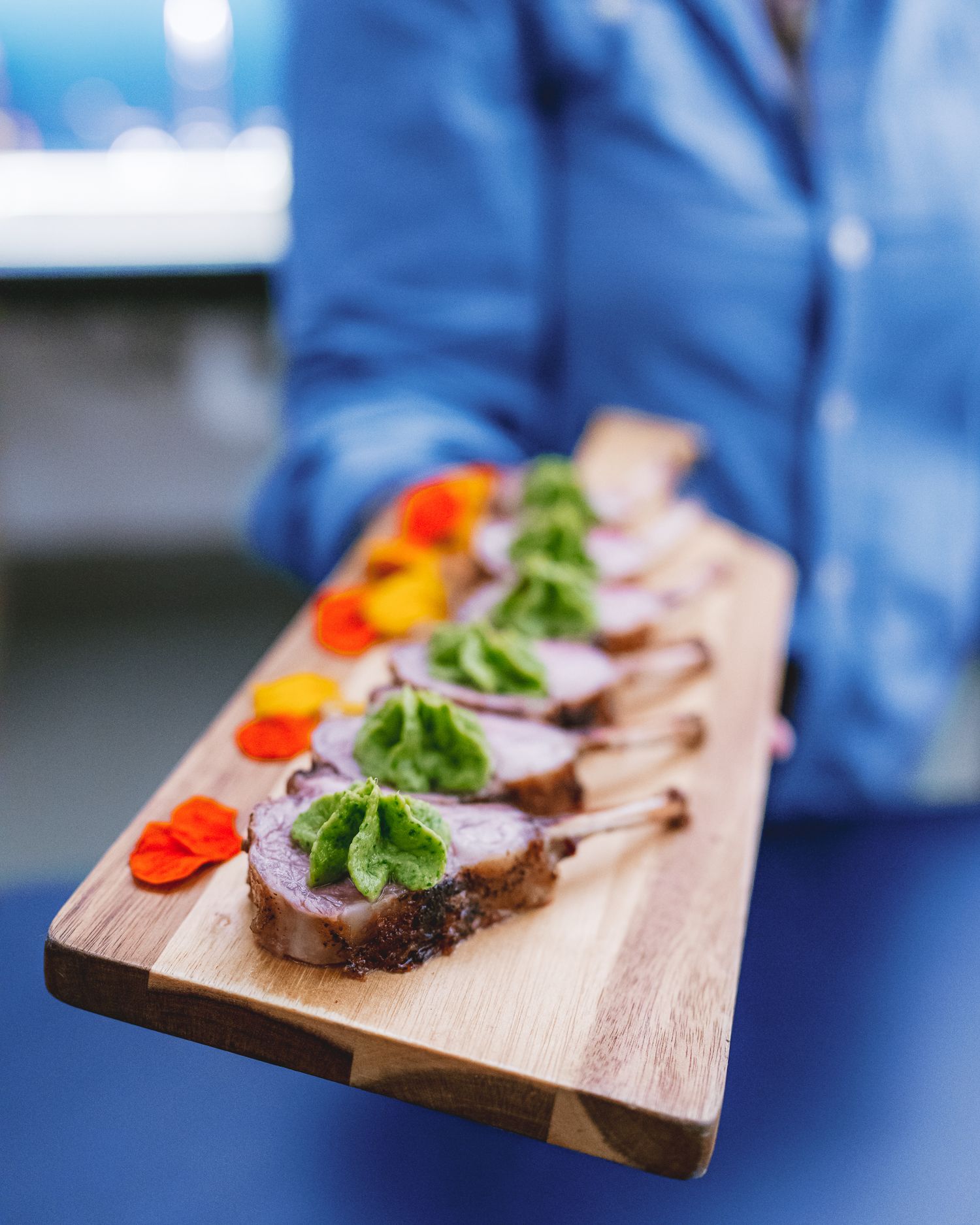 Wooden serving board with sliced meat canapes topped with fresh greens and red tomato garnish served against blue backdrop.