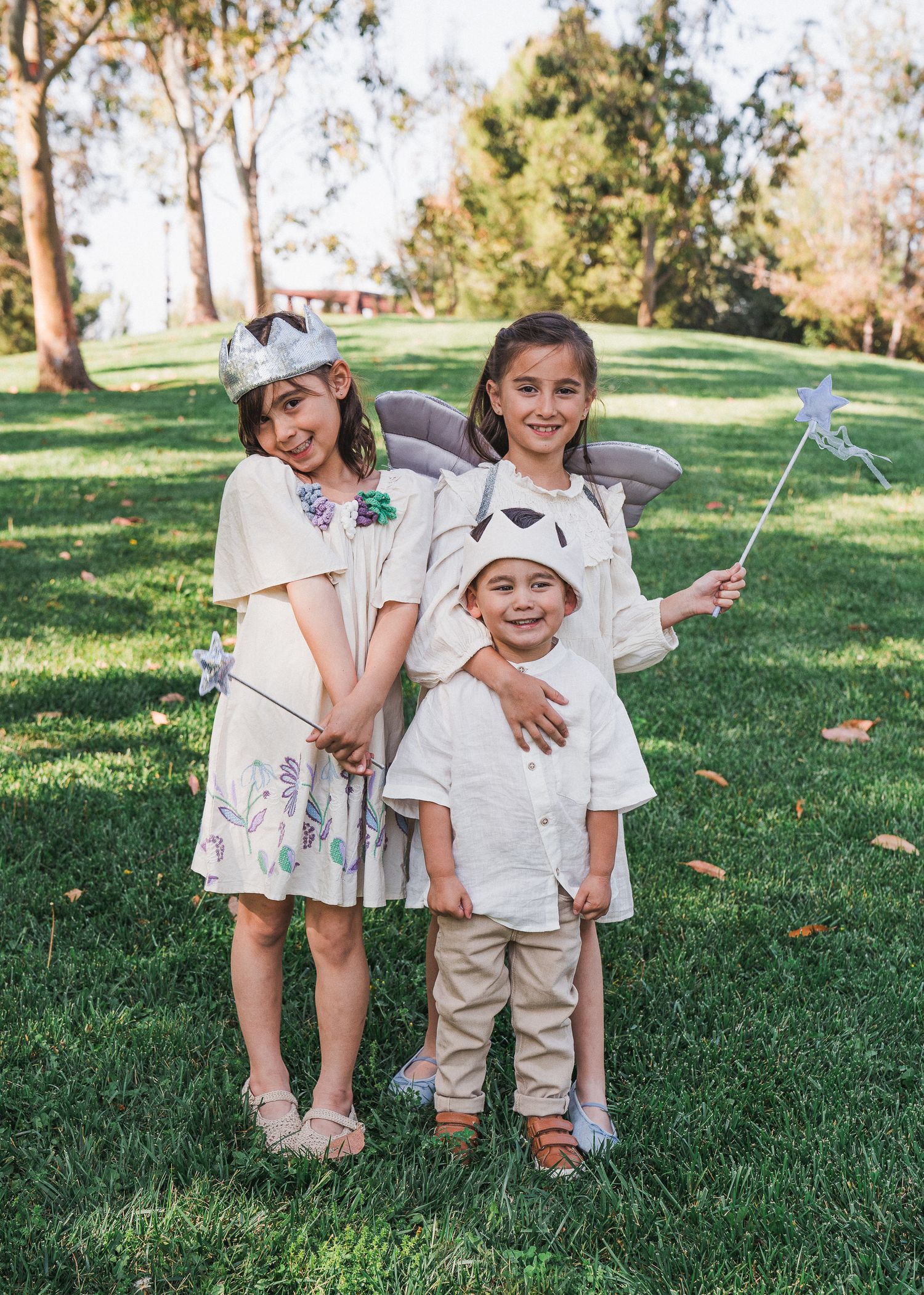 Children wearing white outfits stand together on green grass in a sunny park setting with trees in the background.