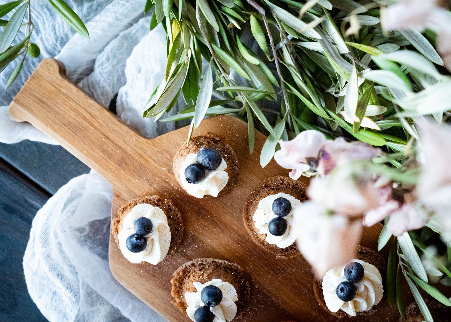 Mini chocolate desserts topped with whipped cream and blueberries served on a wooden board with fresh greenery.