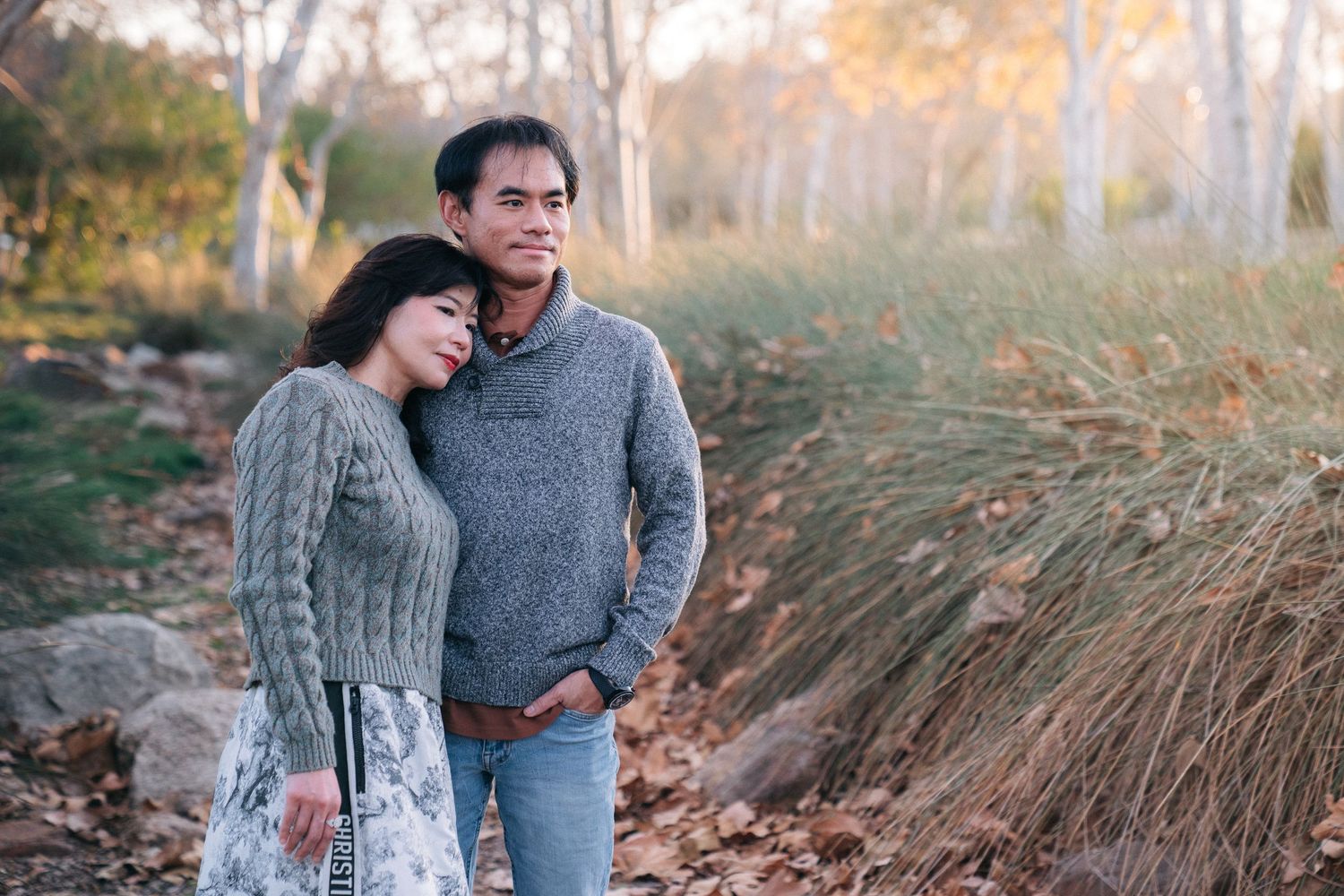 A couple stands together in a romantic autumn scene with tall dried grasses and trees in the background.
