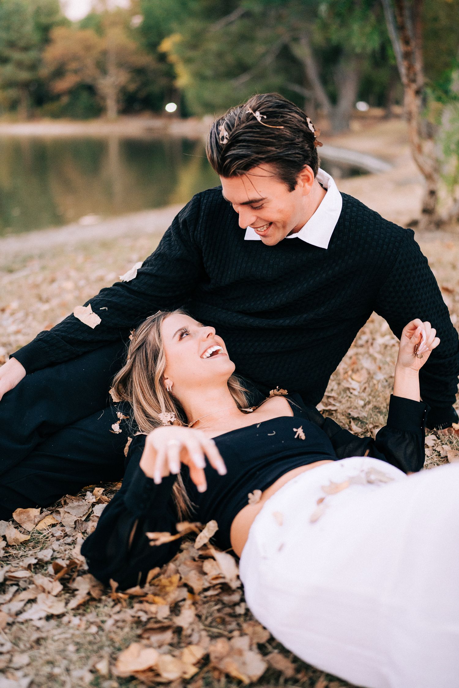 A candid moment of laughter captured between a couple sitting on autumn leaves by a lake.