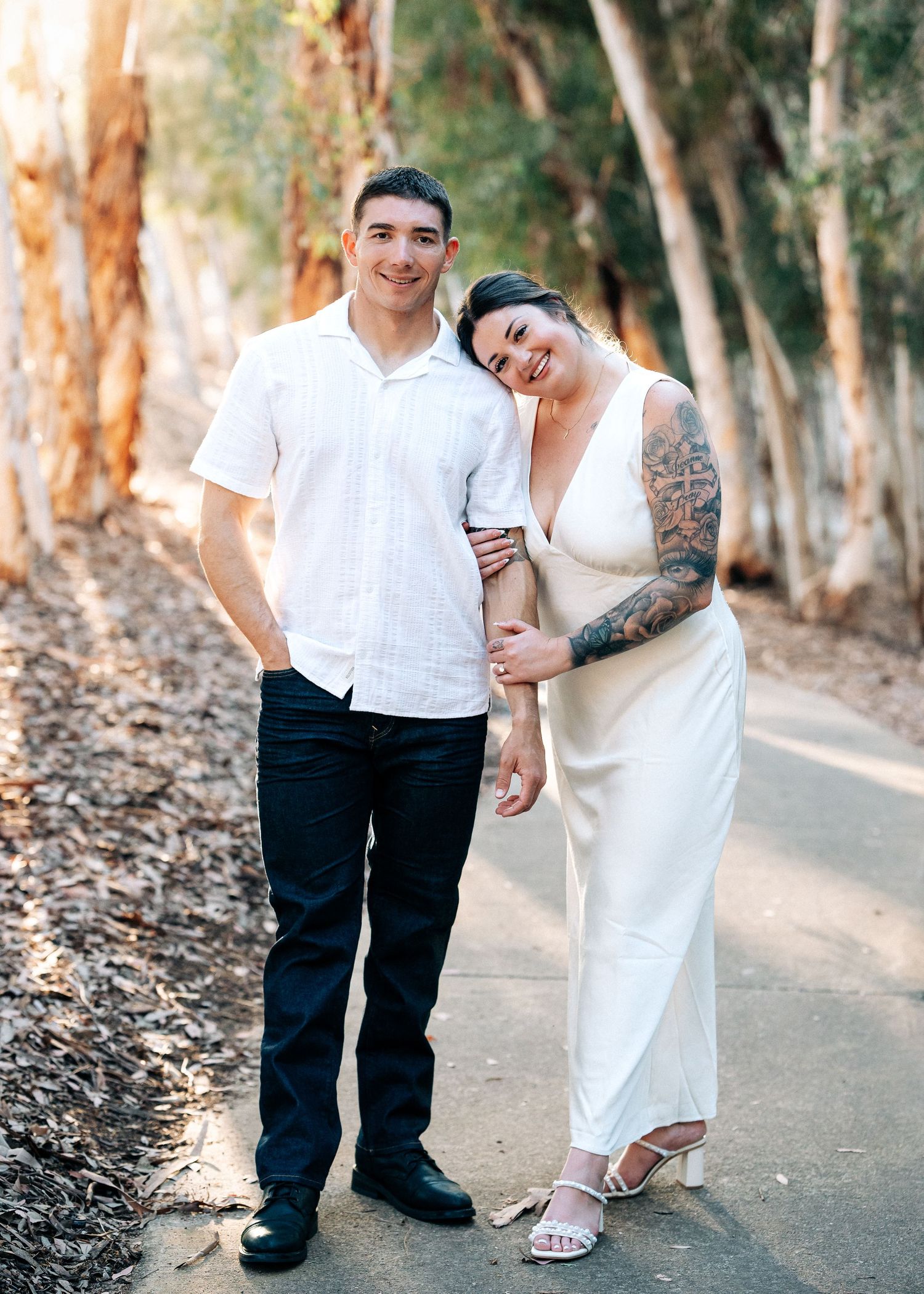 A couple in white outfits poses together on a tree-lined path, with natural sunlight filtering through the eucalyptus trees.