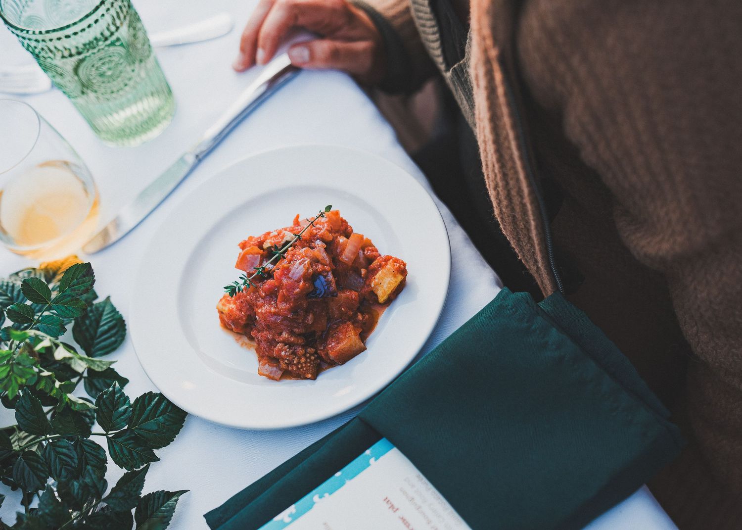 A plate of spicy red tomato-based dish served on a white plate with greenery and drinks in the background.