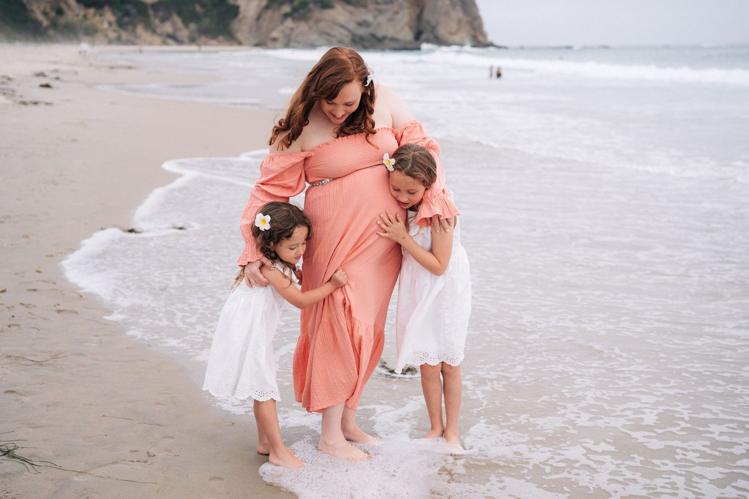 Three people in coral and white dresses share a tender moment together on a misty beach.